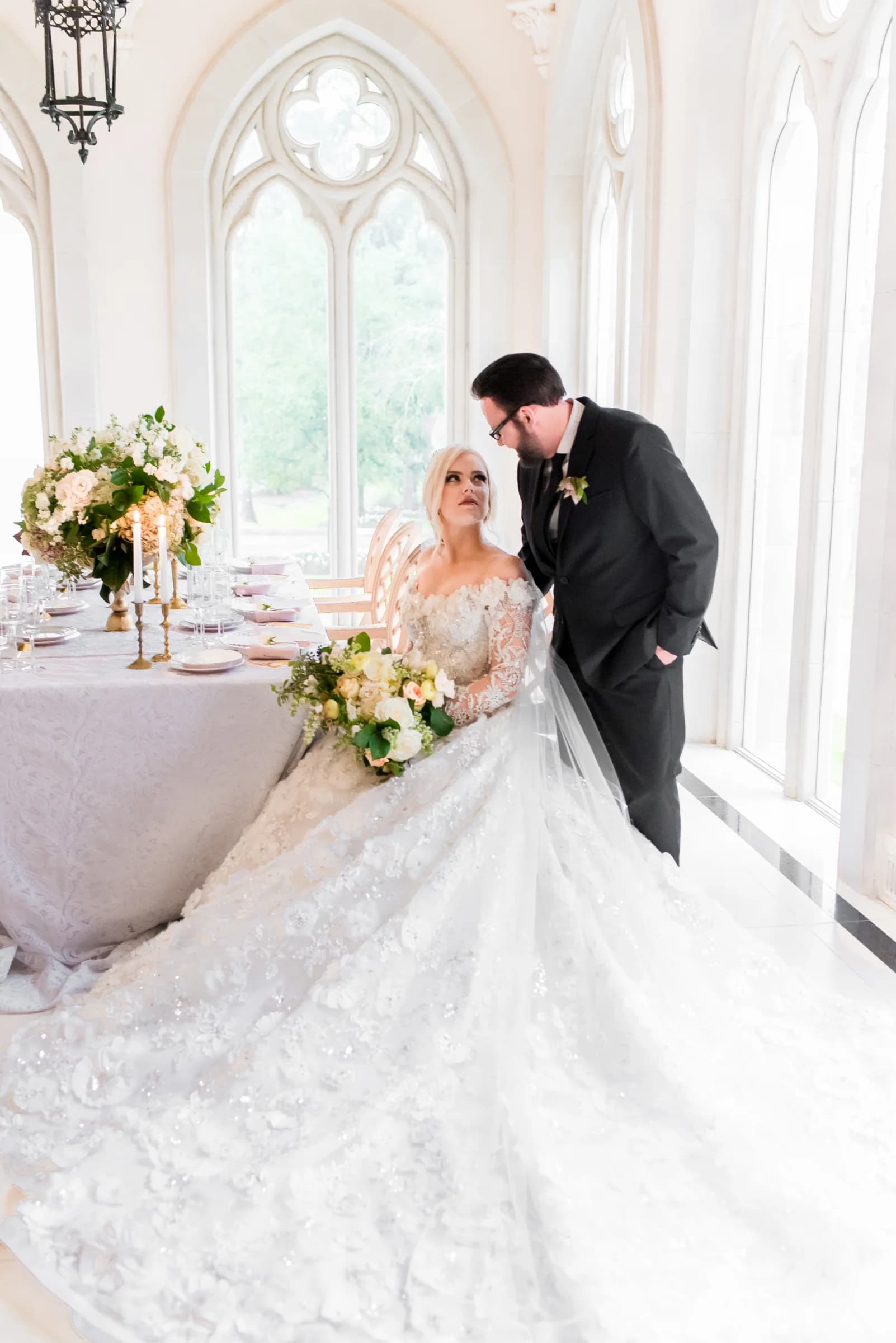 A bride in a stunning gown gazes at her groom in an elegant venue adorned with floral arrangements and soft natural light.
