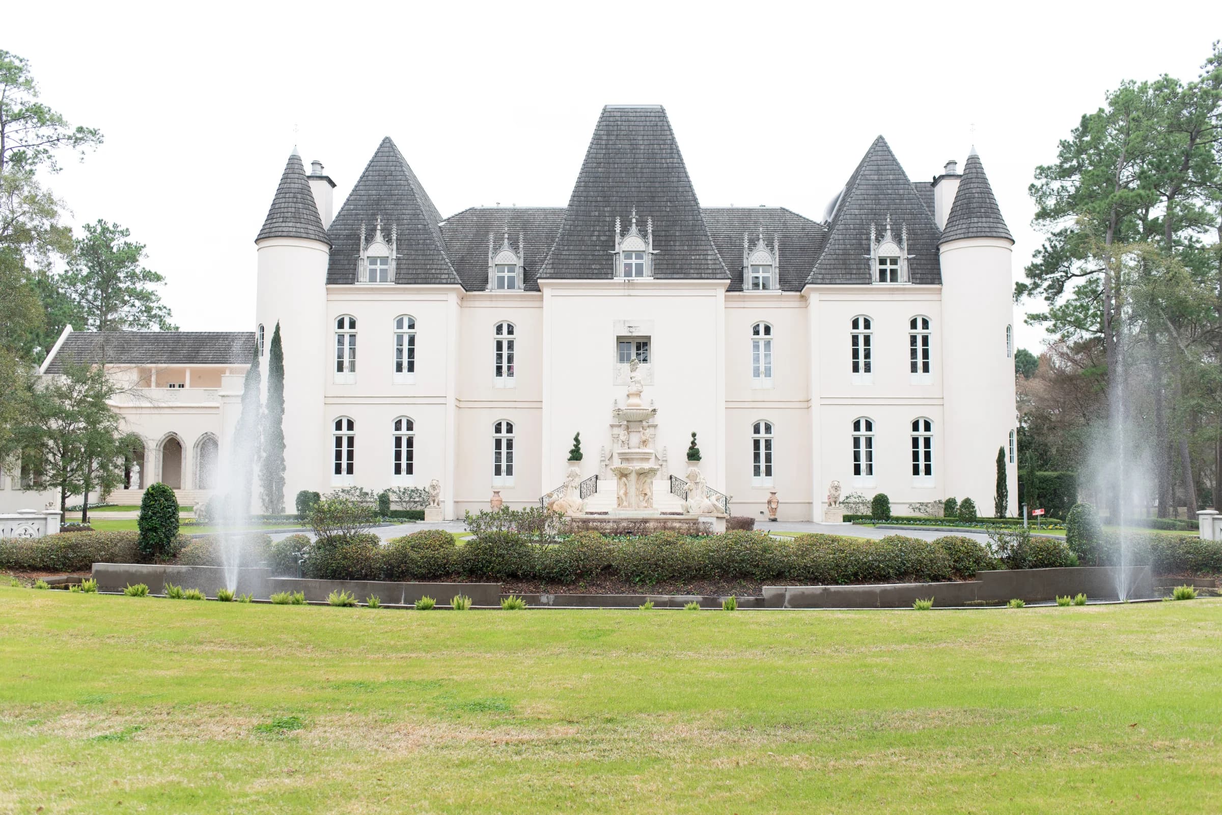 A grand chateau with pointed roofs and a central fountain, surrounded by manicured lawns and lush greenery.