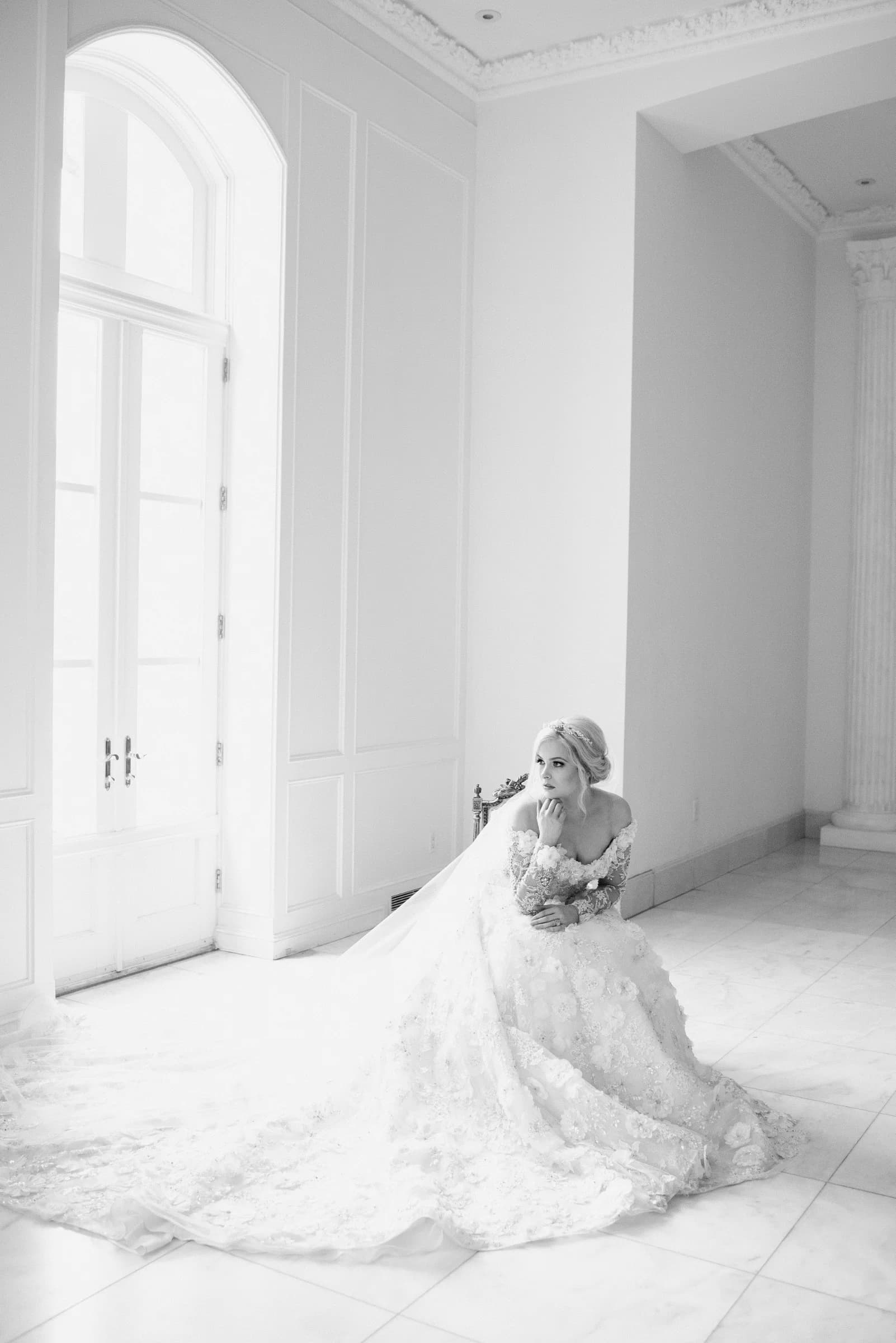 A bride in an elegant gown sits thoughtfully in a bright, minimalist room, surrounded by soft light and white decor.