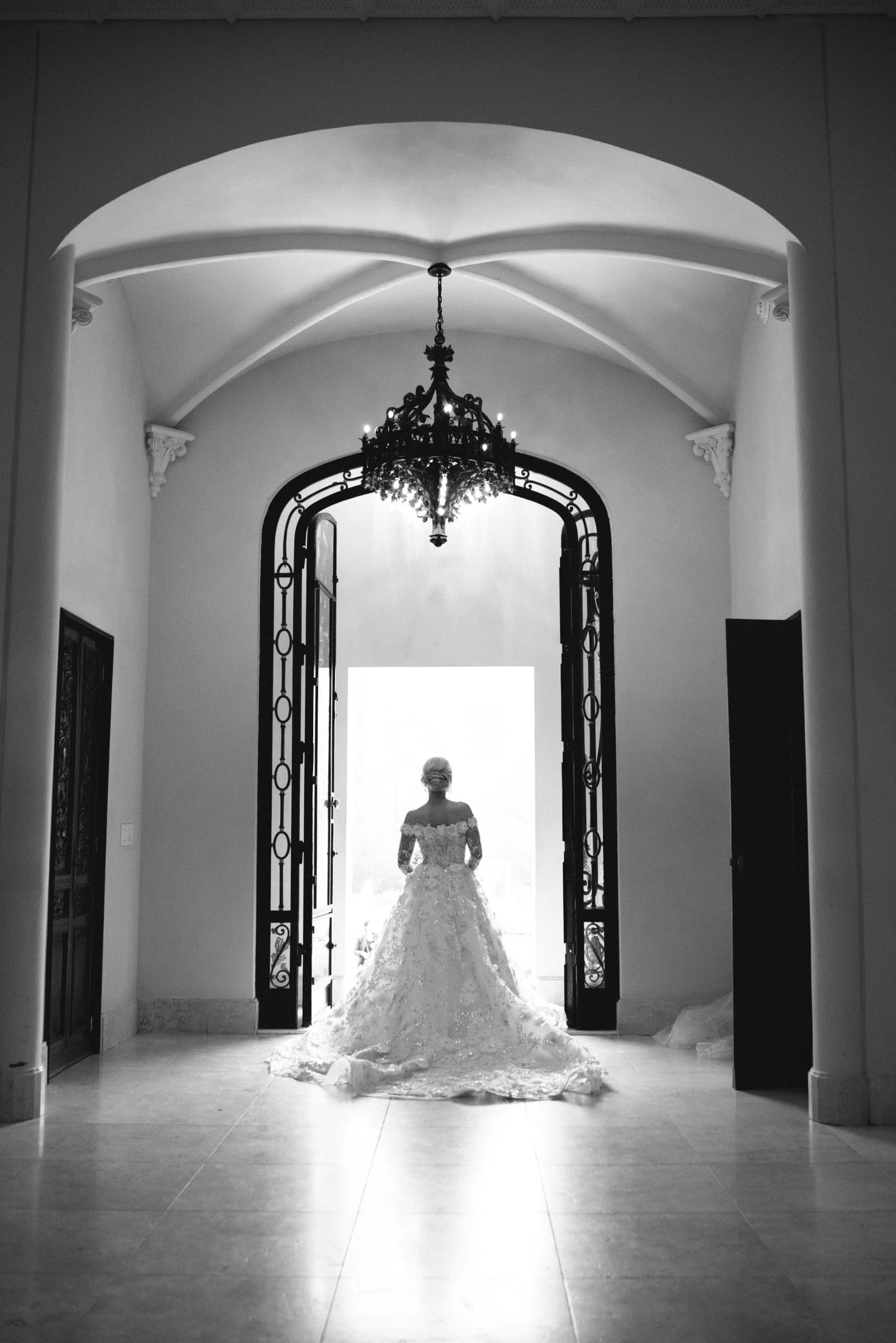 A bride in a detailed gown stands in an elegant doorway, framed by ornate architecture and a chandelier above.