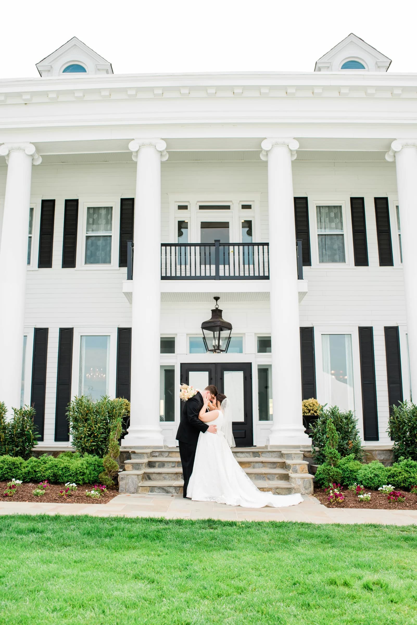 A couple shares a romantic kiss on the steps of a grand white mansion, surrounded by lush greenery and blooming flowers.