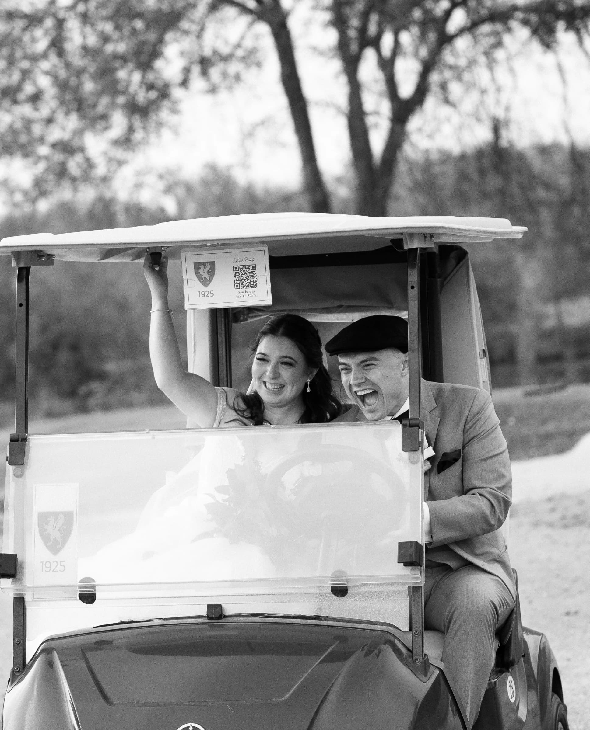 A joyful couple celebrates in a golf cart, with the bride raising her arm and both laughing amidst a scenic backdrop.