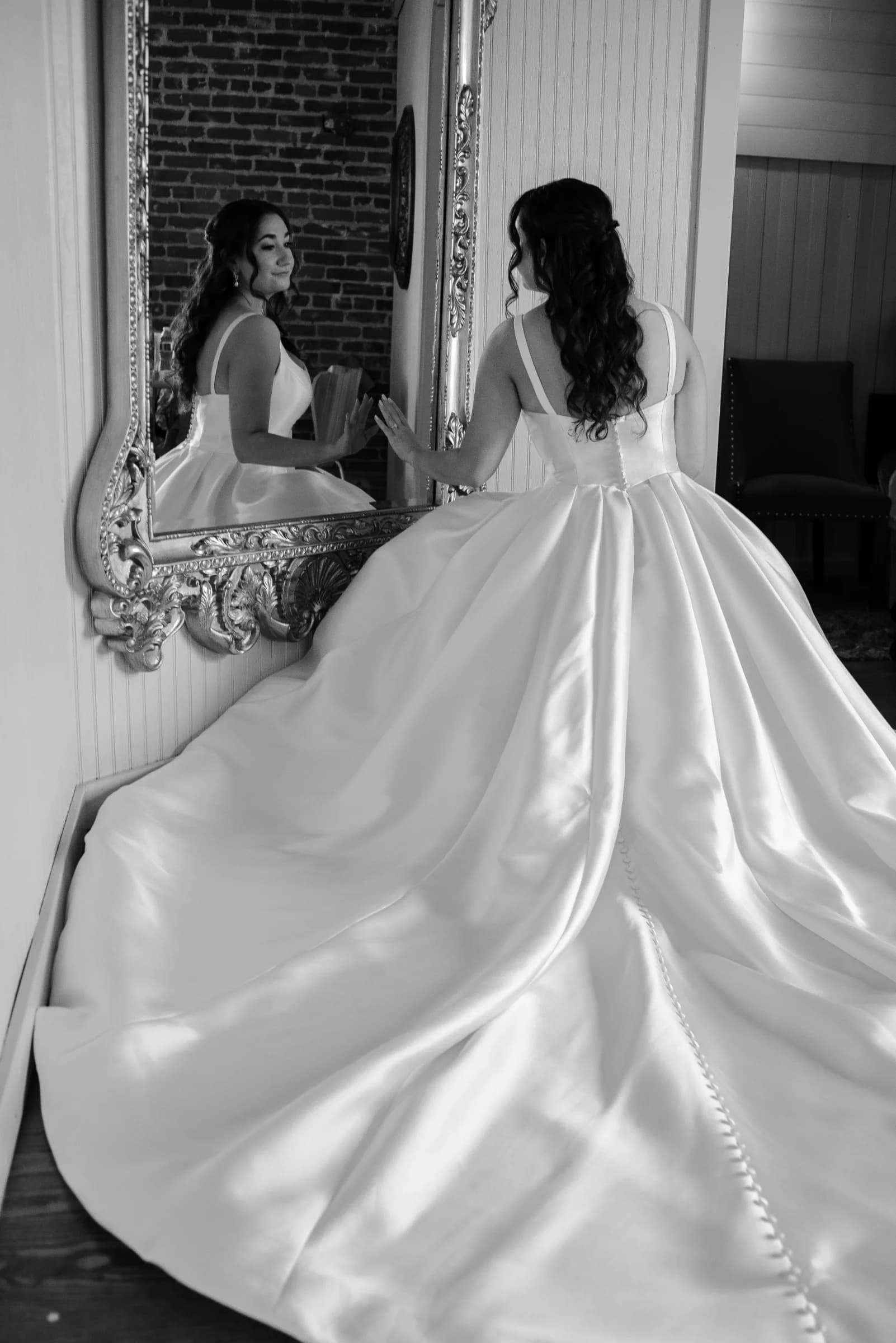 A bride in a flowing white gown admires her reflection in an ornate mirror, exuding elegance and joy.