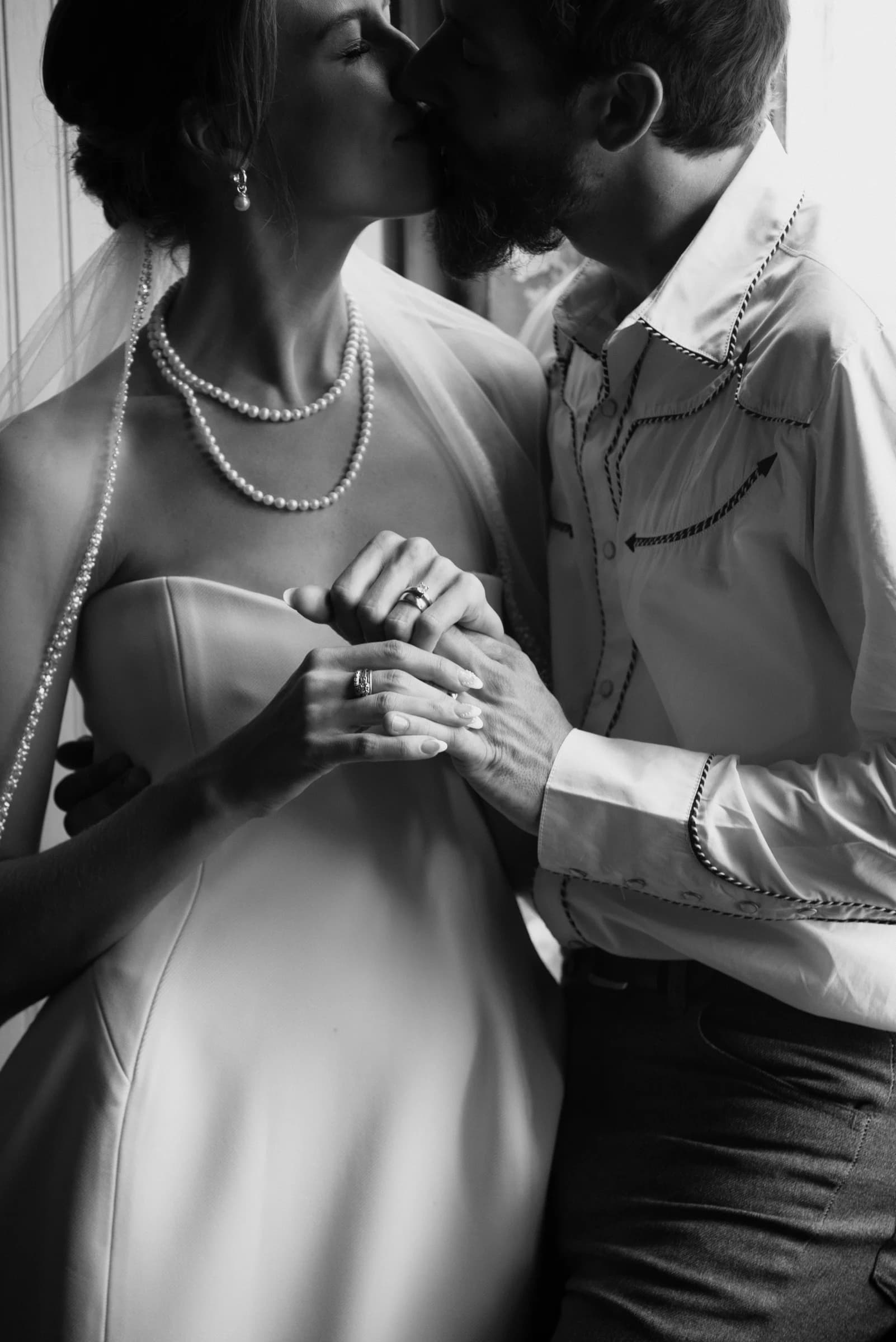 A couple shares a tender kiss, adorned in elegant attire, with their hands intertwined, capturing a moment of intimacy.