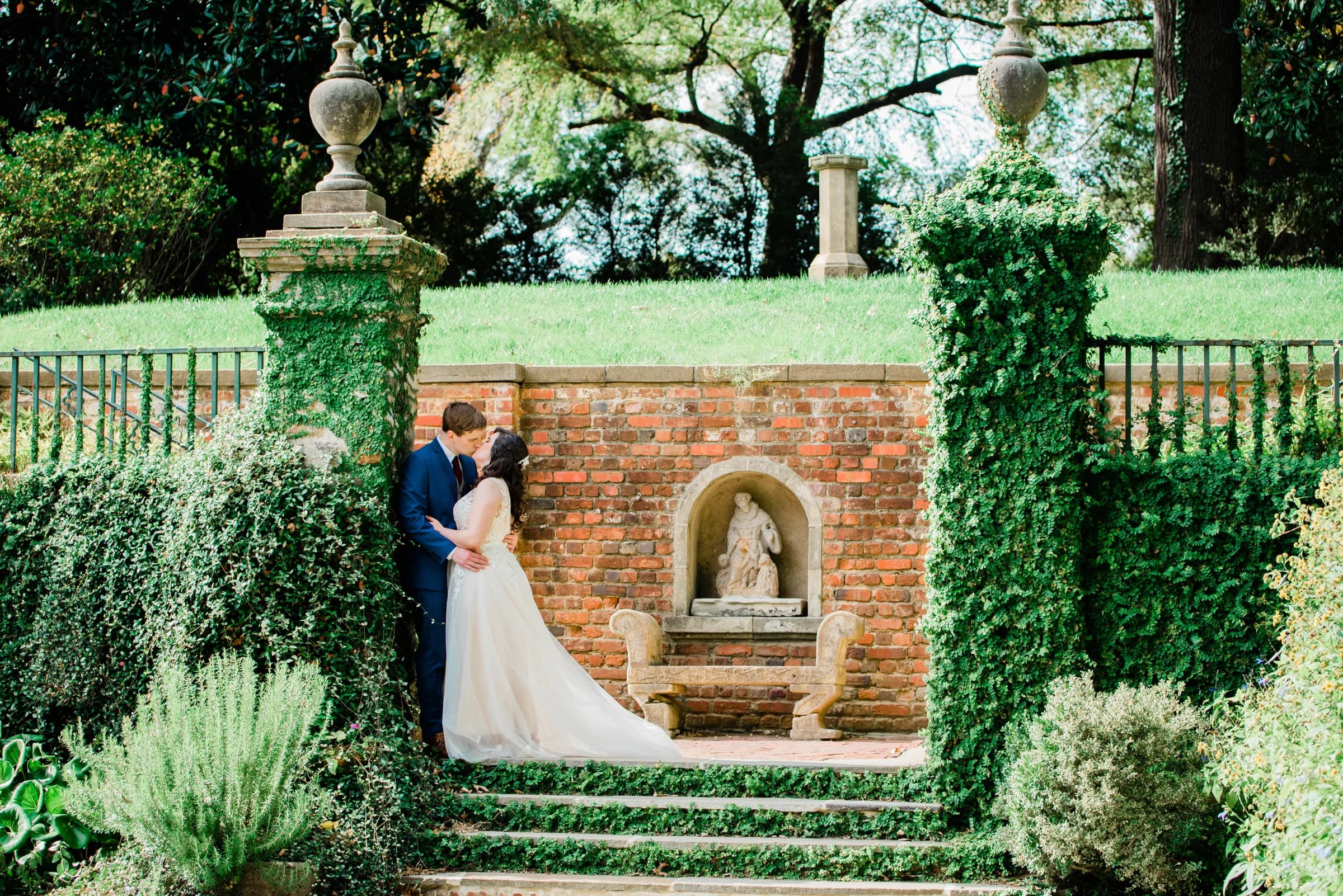 A couple shares a romantic kiss in a lush garden, framed by ivy-covered stone pillars and a serene brick backdrop.