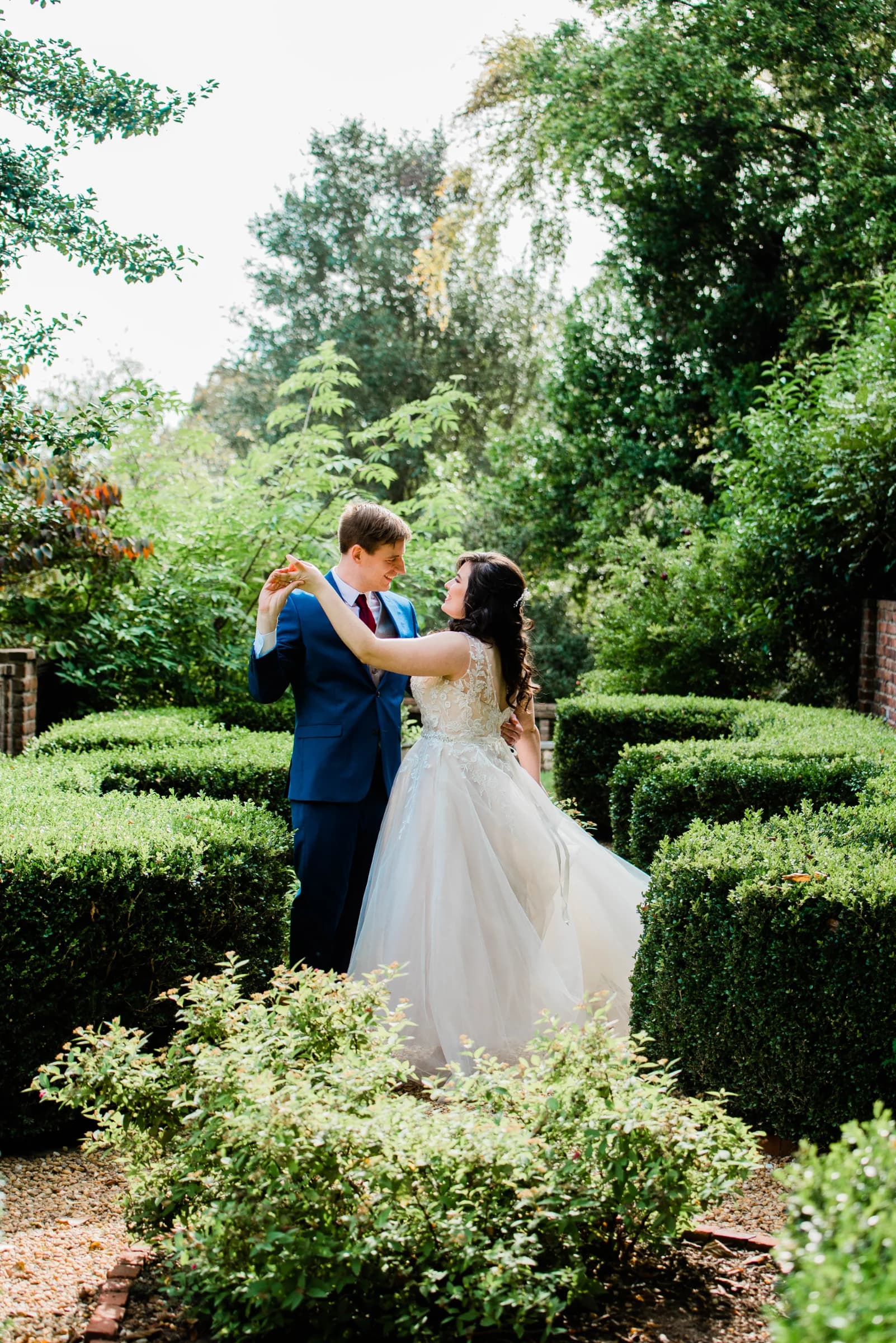A couple dances joyfully in a lush garden, surrounded by neatly trimmed hedges and vibrant greenery.