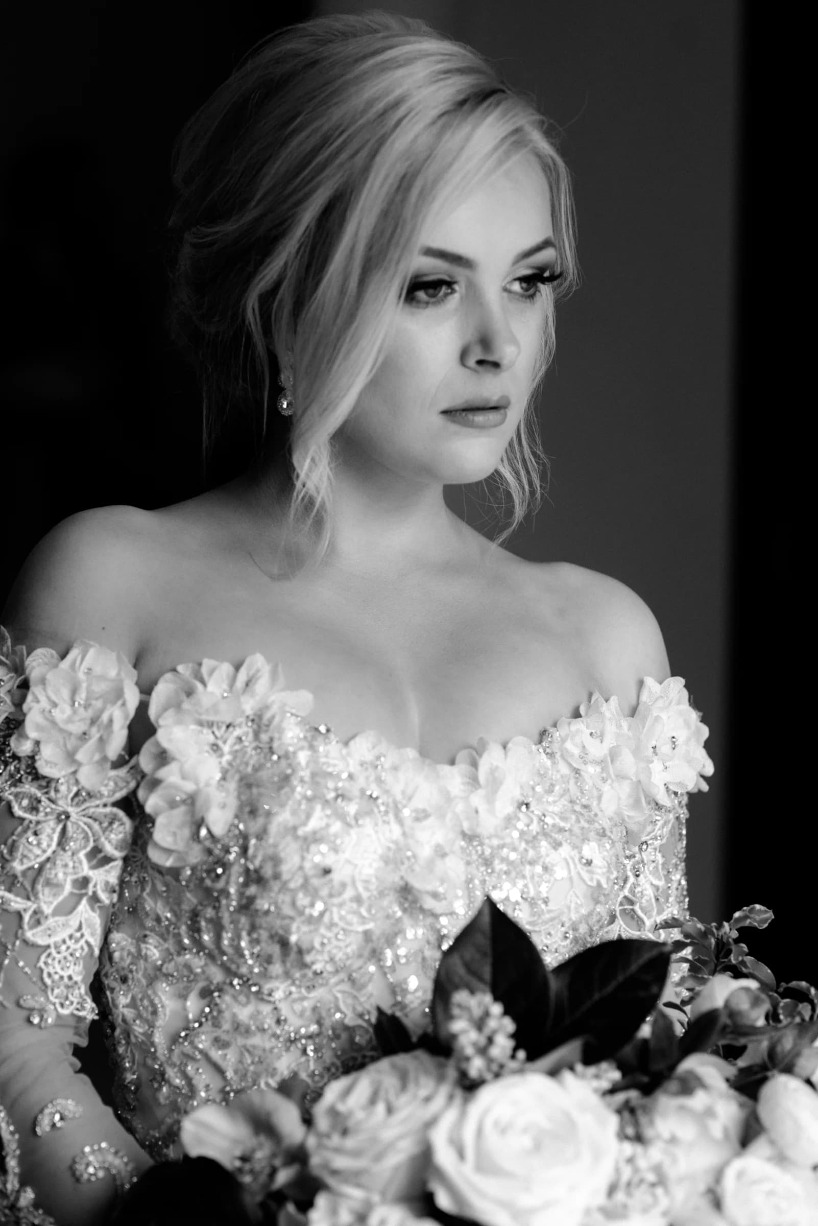 A bride in an intricately beaded gown gazes thoughtfully, holding a bouquet of white roses against a softly lit backdrop.