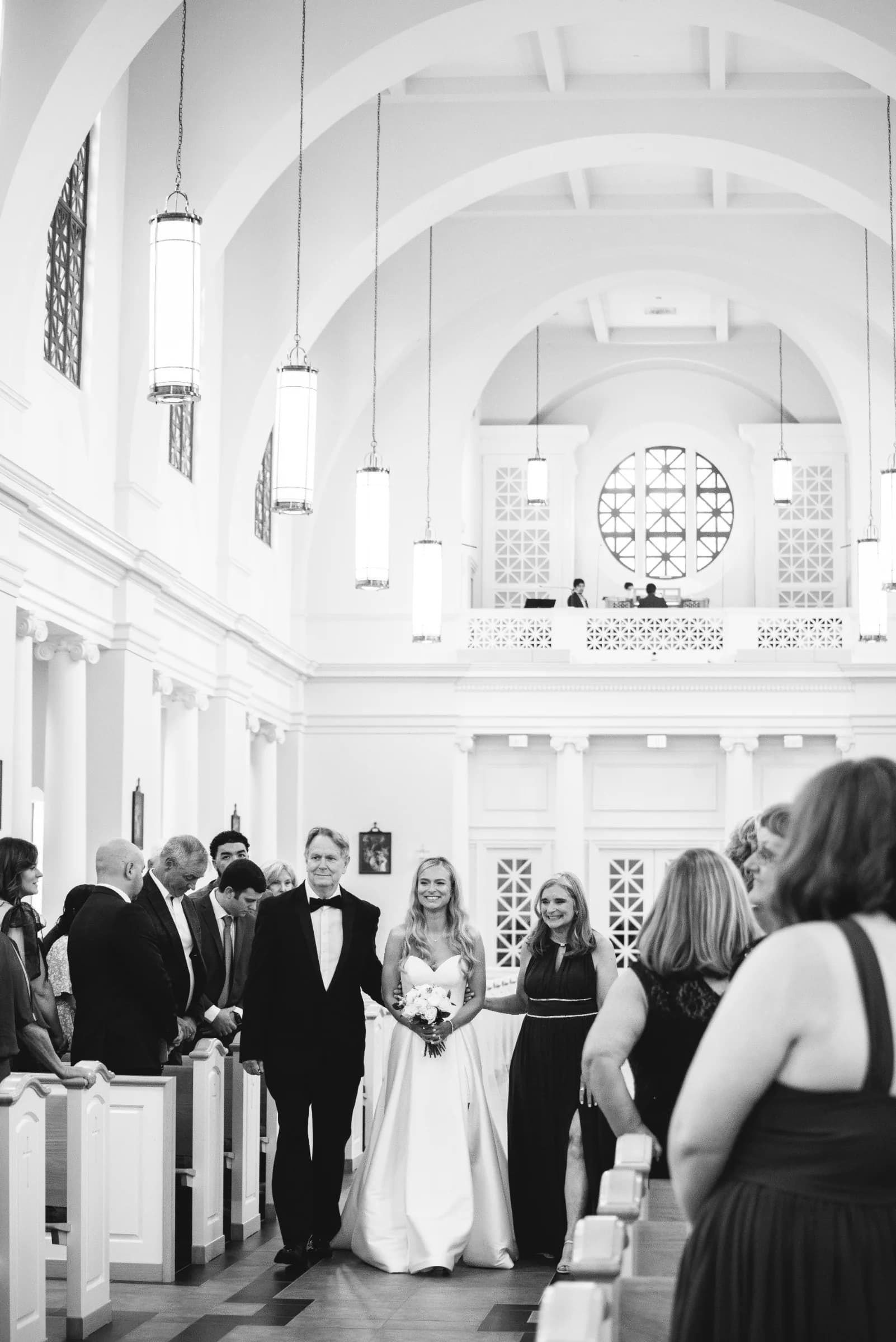 A bride smiles as she walks down the aisle with her father, surrounded by guests in a beautifully lit, elegant venue.