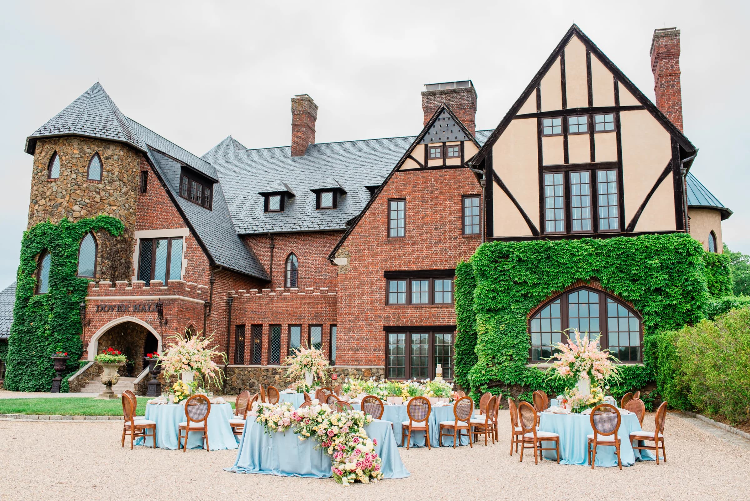 An elegant outdoor dining setup with floral arrangements in front of a charming brick mansion, surrounded by lush greenery.