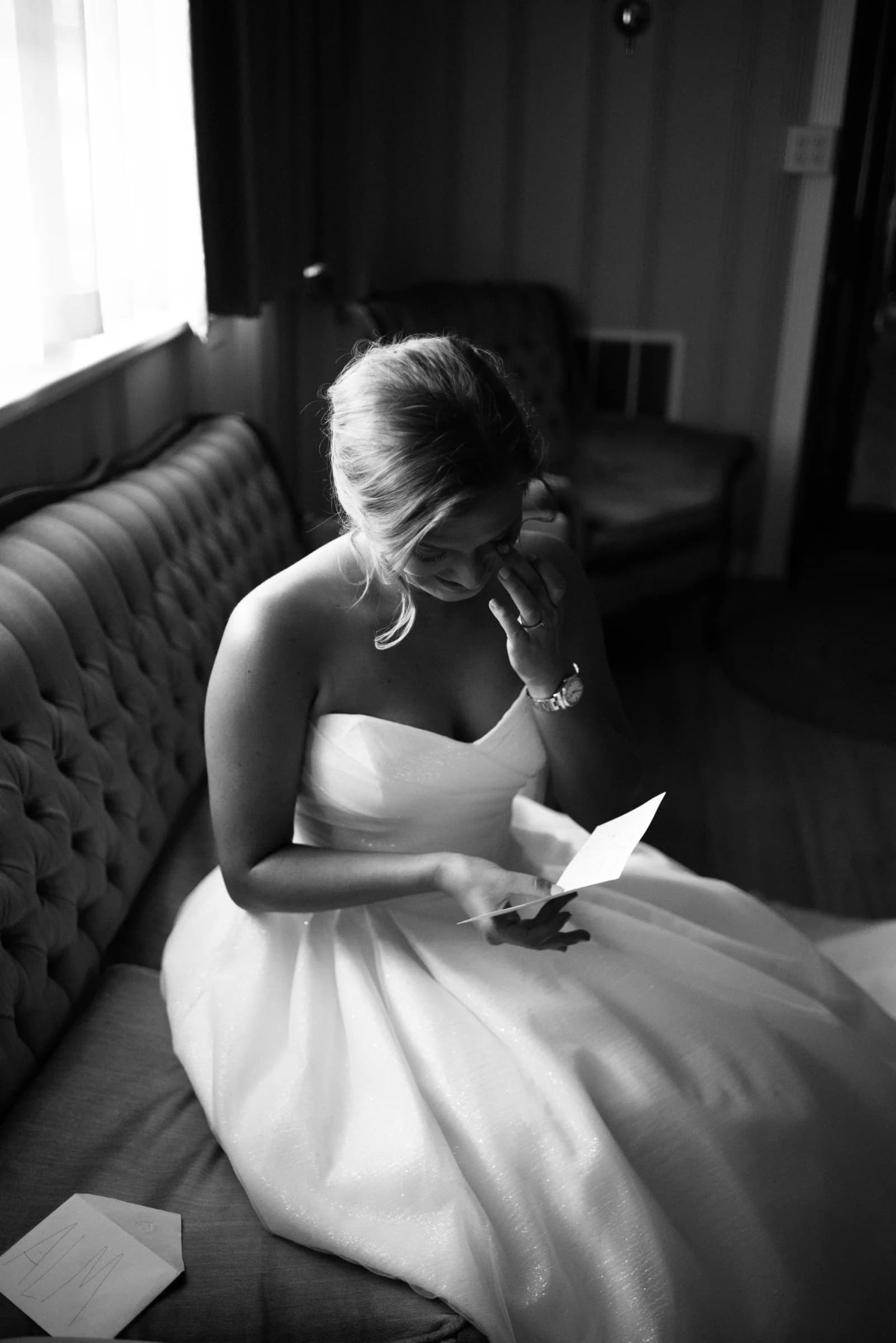 A bride sits on a vintage couch, reading a note with a thoughtful expression, illuminated by soft, natural light.