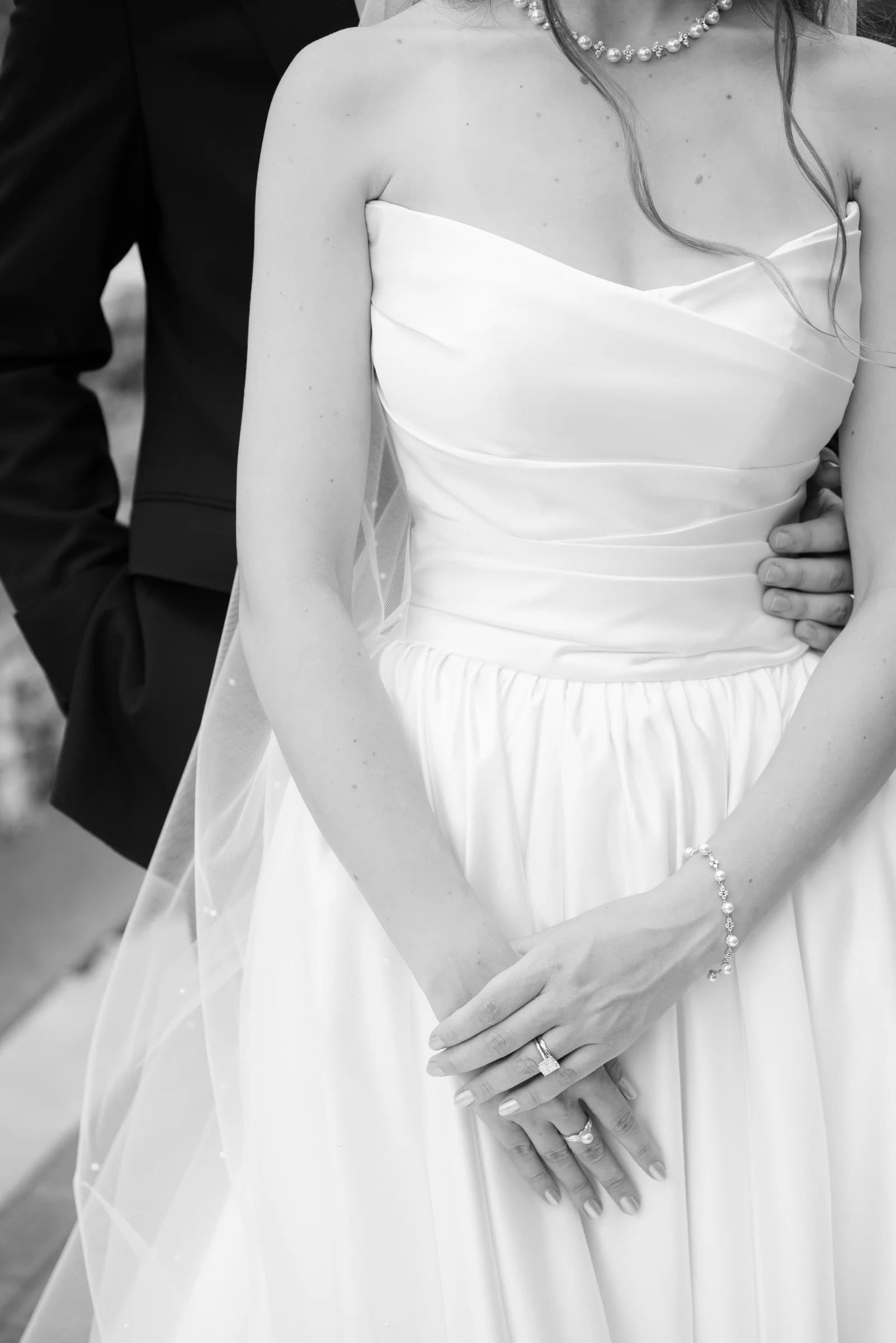 A bride in a strapless white gown holds her hands together, adorned with a bracelet and rings, while a groom's hand rests on her waist.