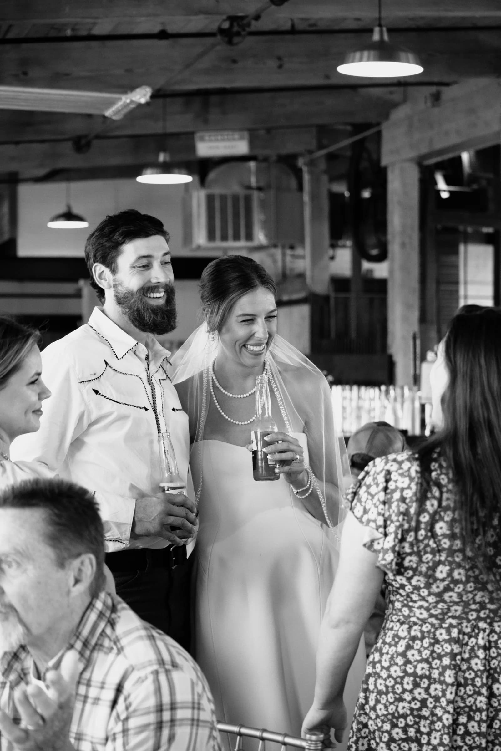 A joyful couple shares a laugh at their wedding reception, surrounded by friends in a rustic venue.