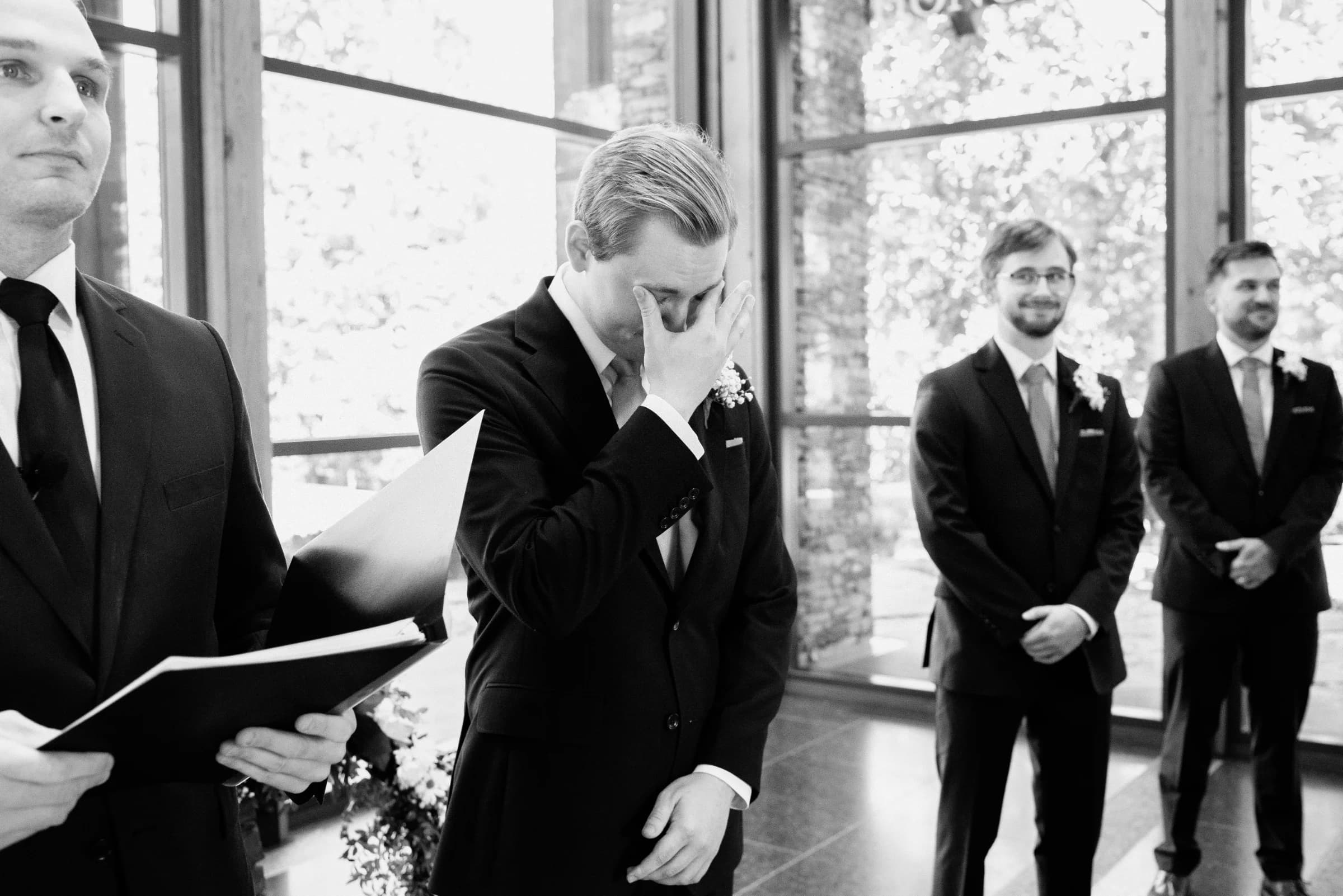 A groom wipes away tears during a heartfelt moment at a wedding ceremony, surrounded by his supportive groomsmen.