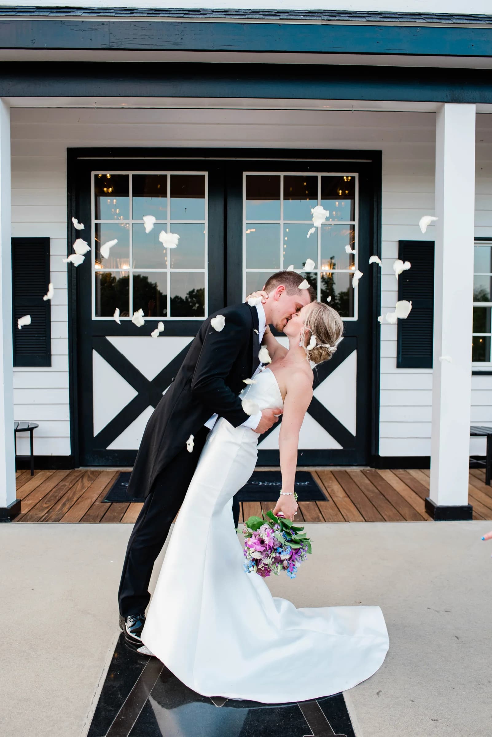 A couple shares a romantic kiss surrounded by falling rose petals outside a charming venue, exuding joy and celebration.