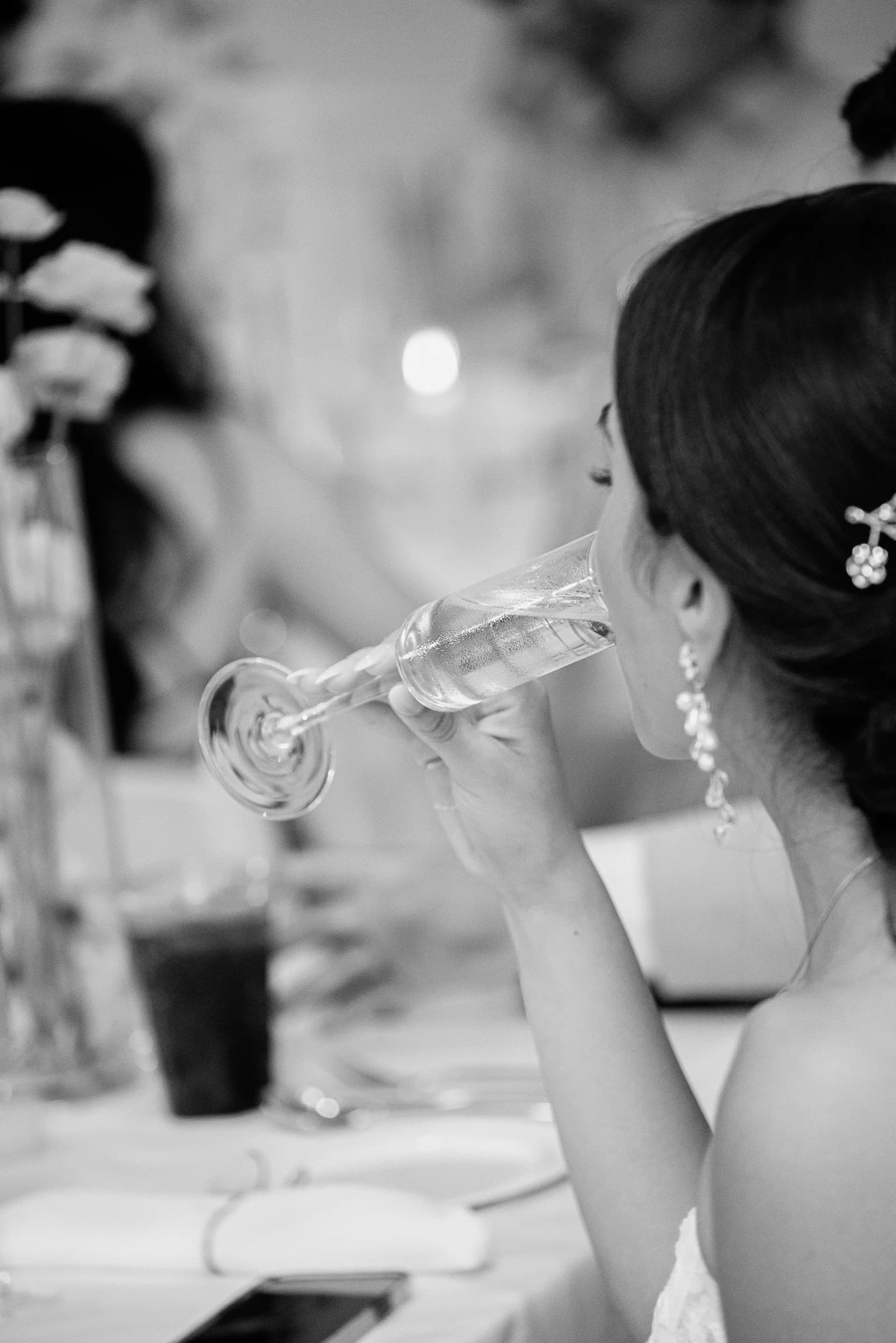 A woman in elegant attire sips from a champagne flute at a beautifully set table, surrounded by soft, blurred decorations.