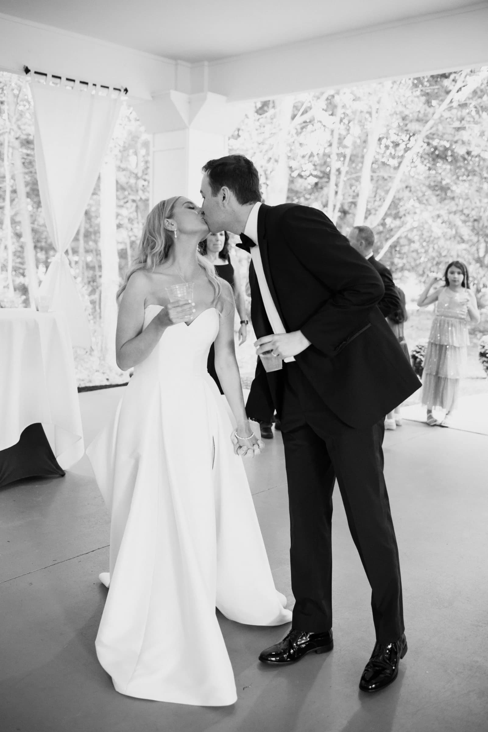 A couple shares a kiss while holding hands, surrounded by guests in a bright, elegant setting.