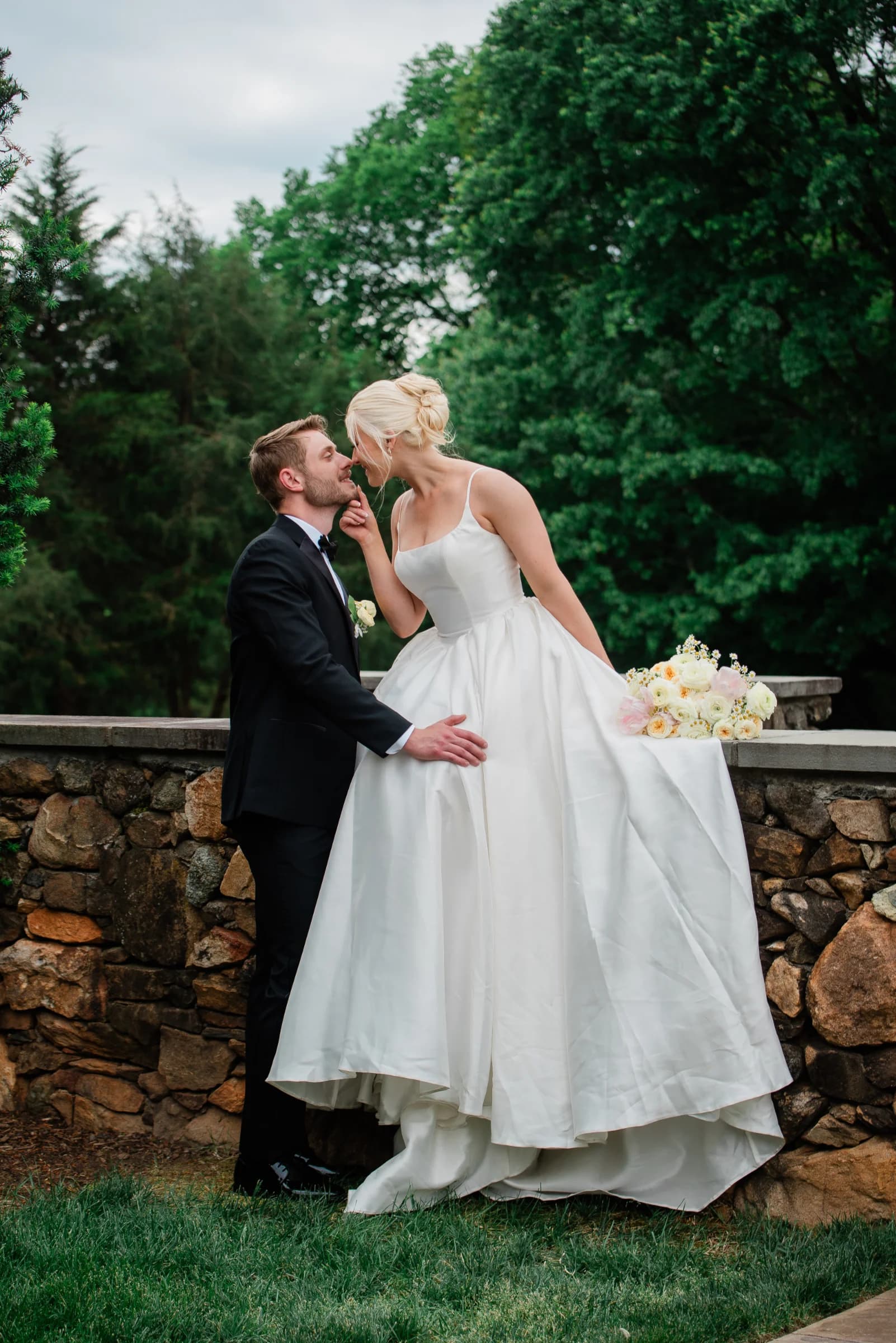 A bride in a flowing white gown playfully touches her groom's face, surrounded by lush greenery and a stone wall.