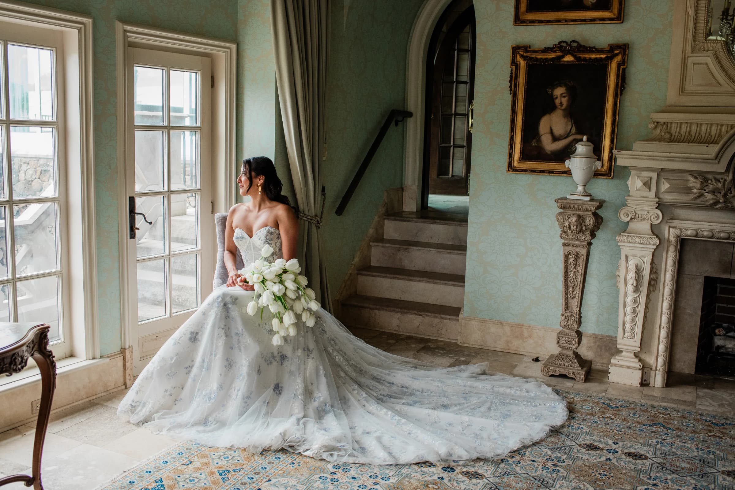 A bride in a stunning gown sits by a window, holding a bouquet of tulips, surrounded by elegant decor and soft natural light.