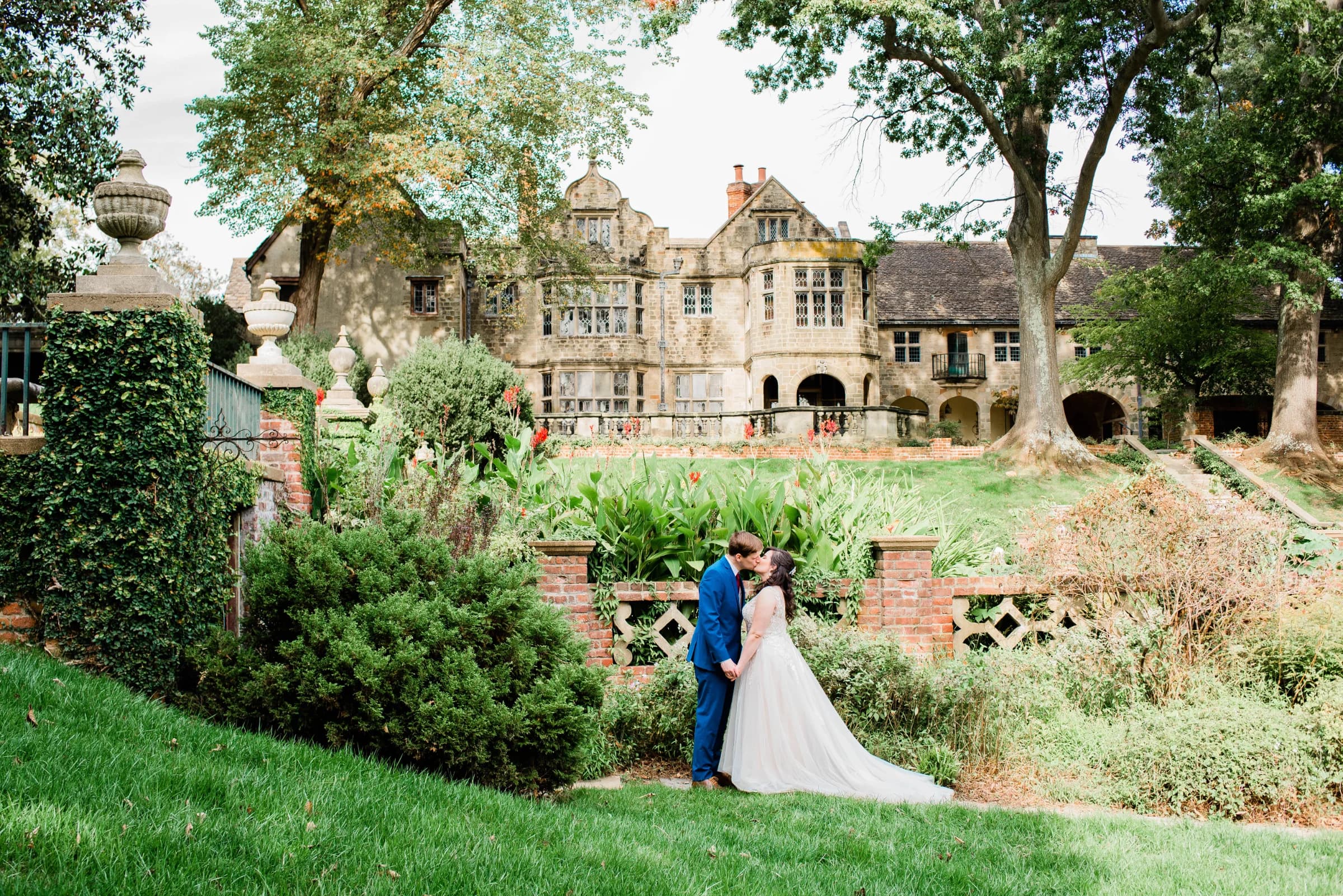 A couple shares a romantic kiss in a lush garden, with a grand historic mansion in the background.