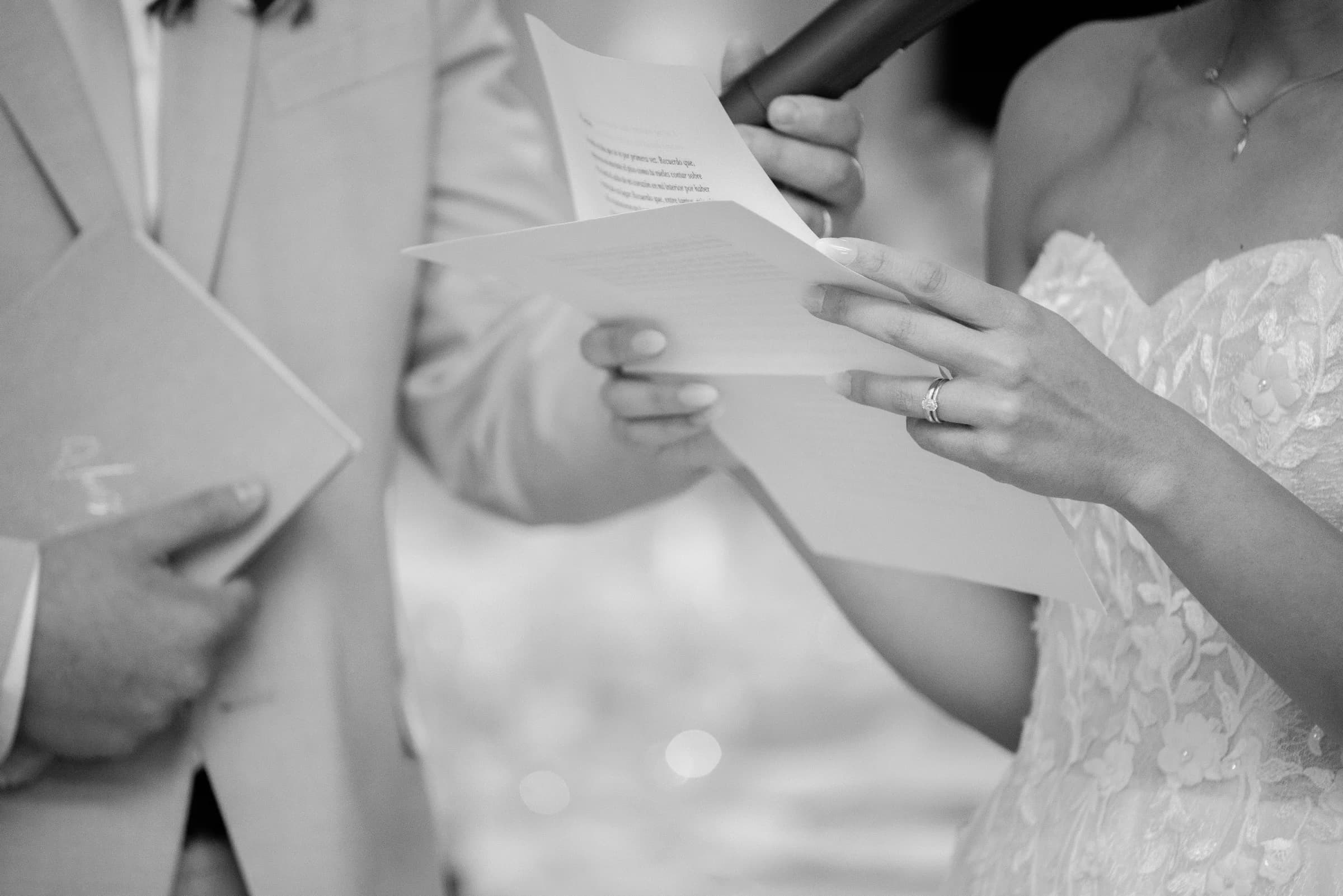 A bride reads vows while holding a microphone, her hand adorned with a ring, as a groom listens attentively.