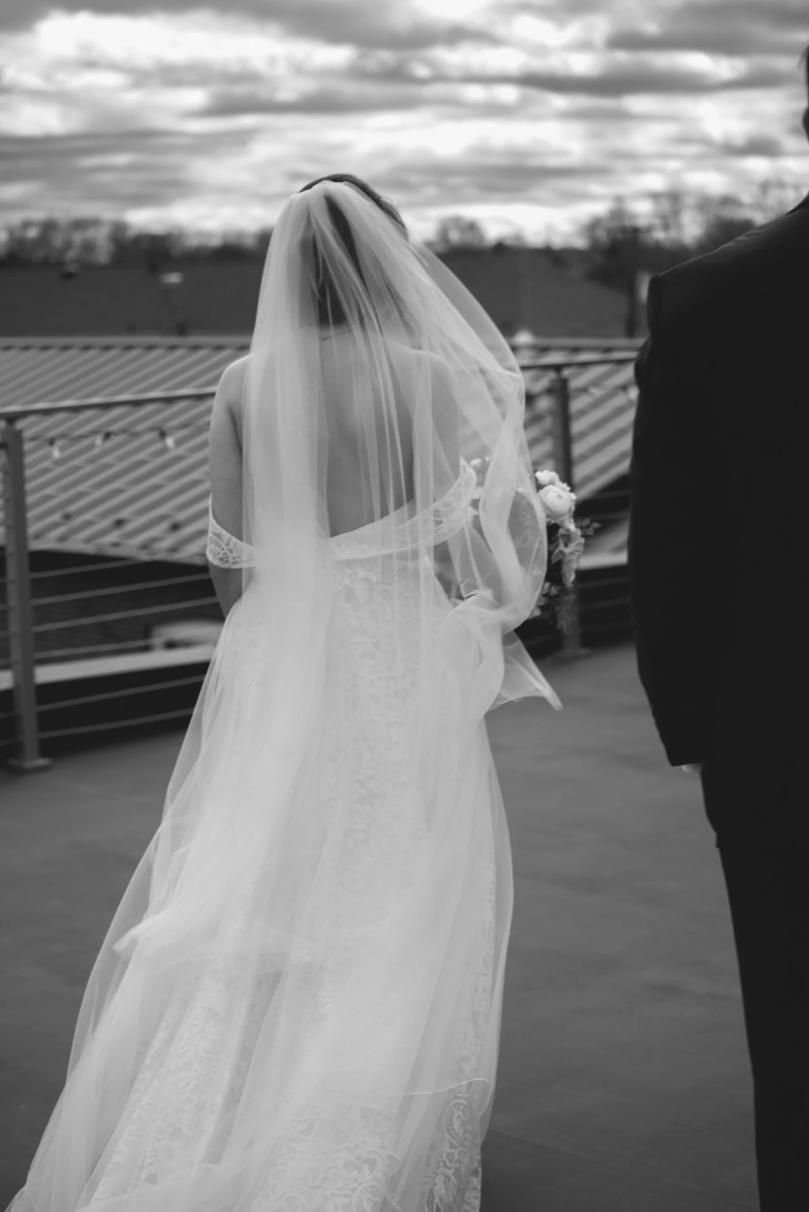 A bride in a flowing white gown and veil stands on a rooftop, gazing into the distance beside a suited figure.