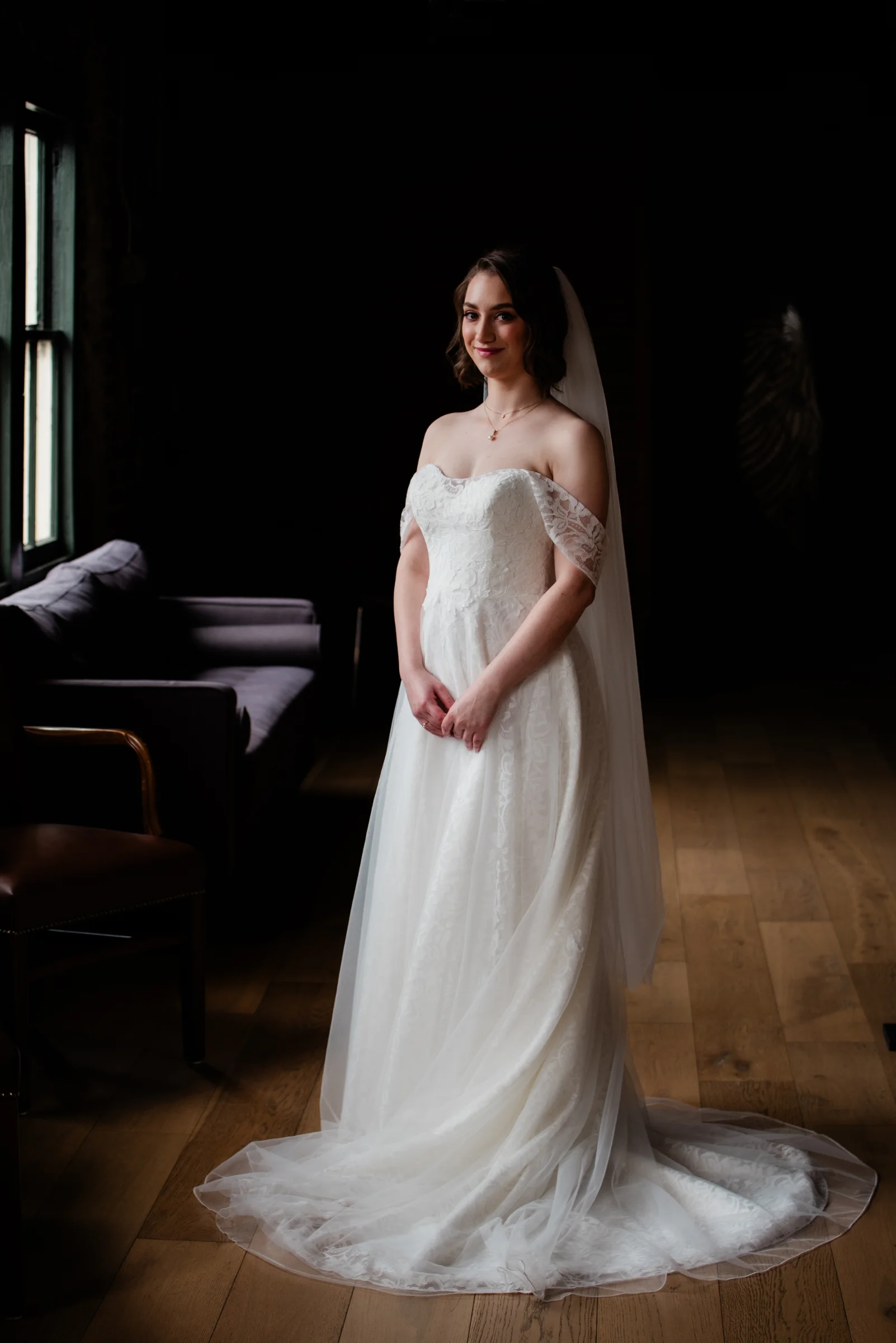A bride in a delicate off-the-shoulder gown stands gracefully in a dimly lit room, exuding elegance and anticipation.