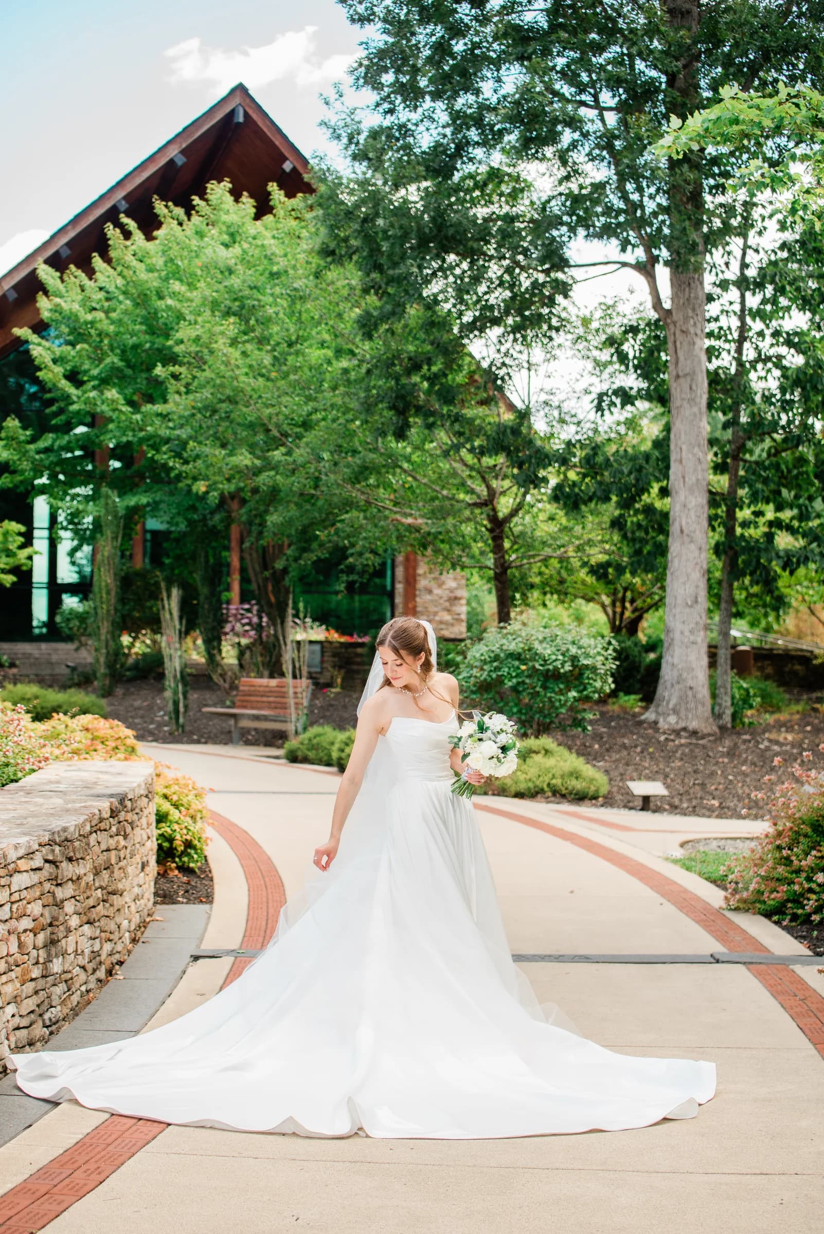 A bride in a flowing white gown gracefully walks along a garden path, holding a bouquet amidst lush greenery and a rustic backdrop.