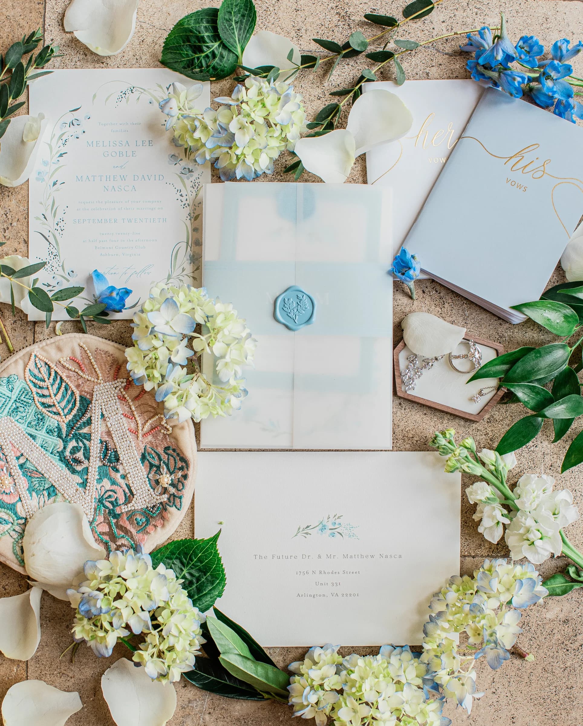 Elegant wedding invitations and vow books surrounded by delicate flowers and greenery on a textured surface.