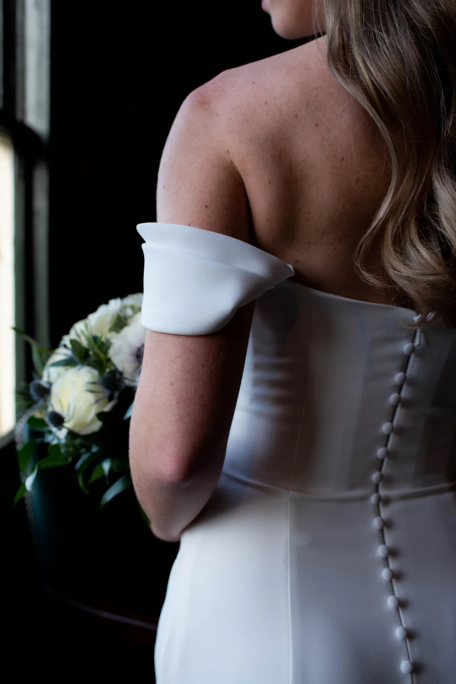 A bride in a sleek white gown holds a bouquet, her back turned to the camera, with soft light illuminating her silhouette.