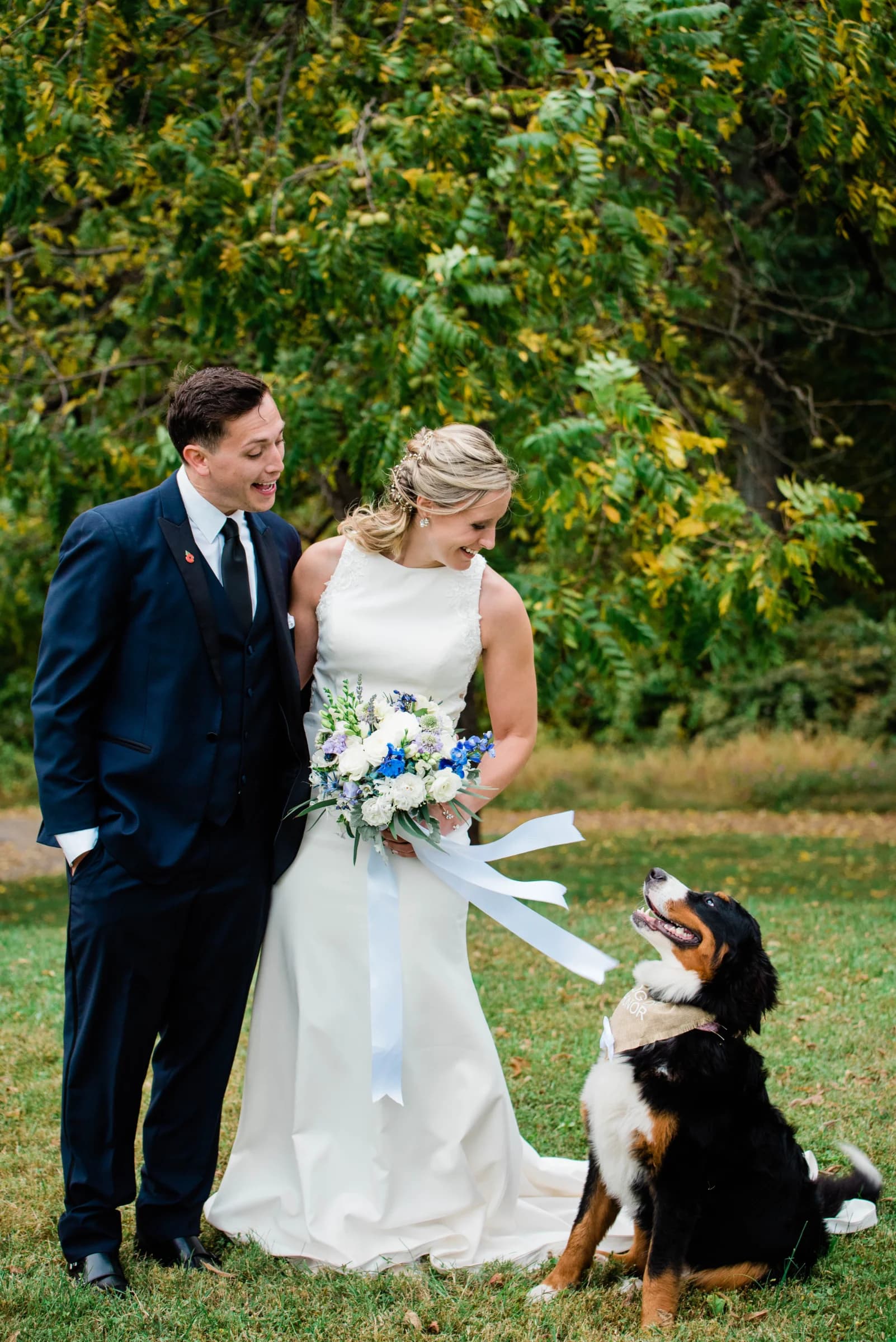 A joyful couple in wedding attire smiles at each other while their dog sits attentively beside them in a lush green setting.