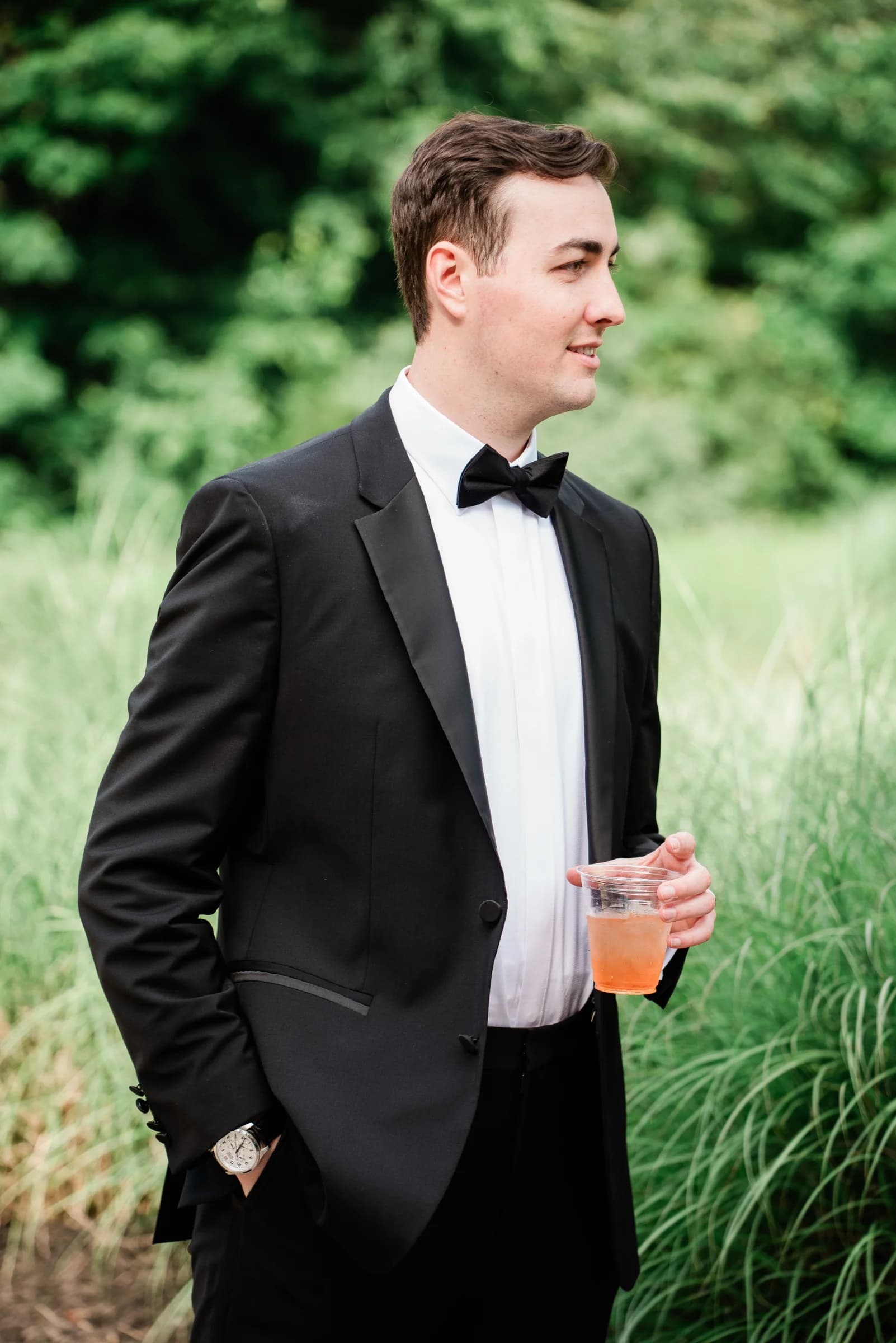 A man in a tuxedo holds a drink, standing confidently among lush green grass.