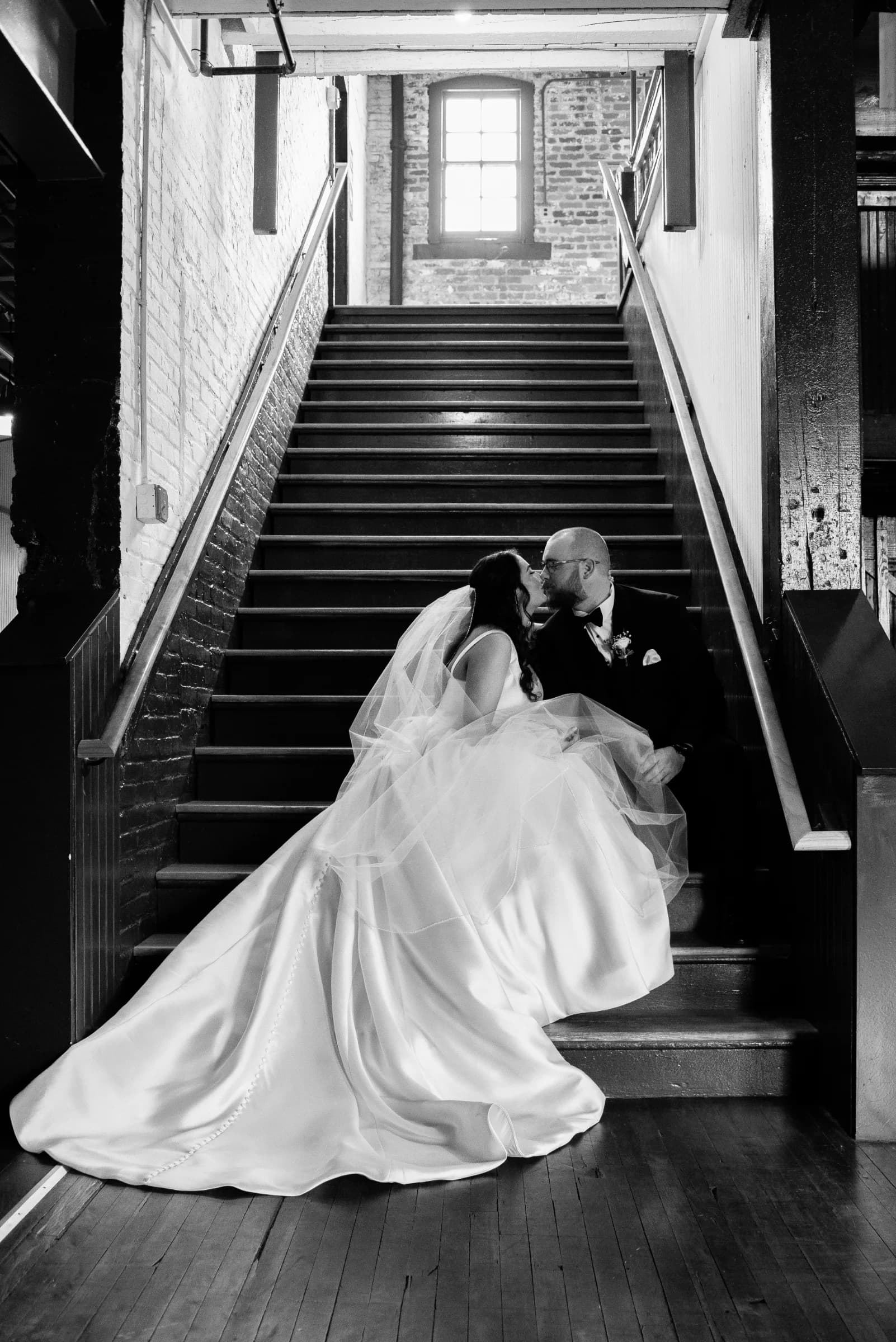 A couple shares a tender kiss on a staircase, surrounded by warm light and rustic brick walls, exuding romance and intimacy.