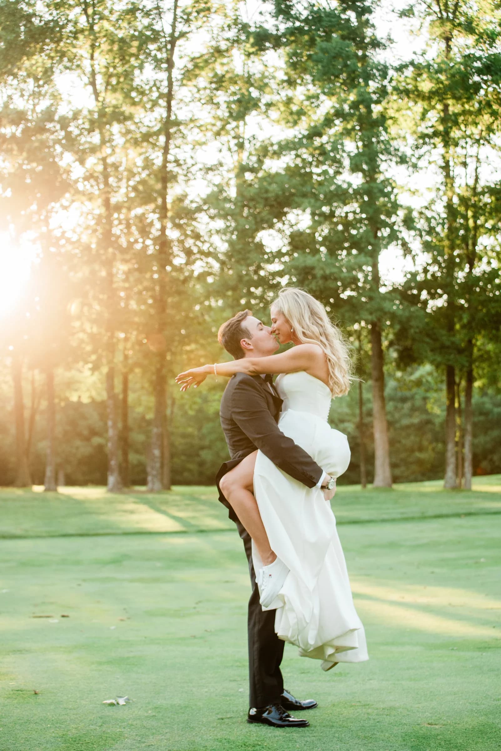 A joyful couple shares a kiss as the groom lifts the bride in a sunlit, lush green setting.
