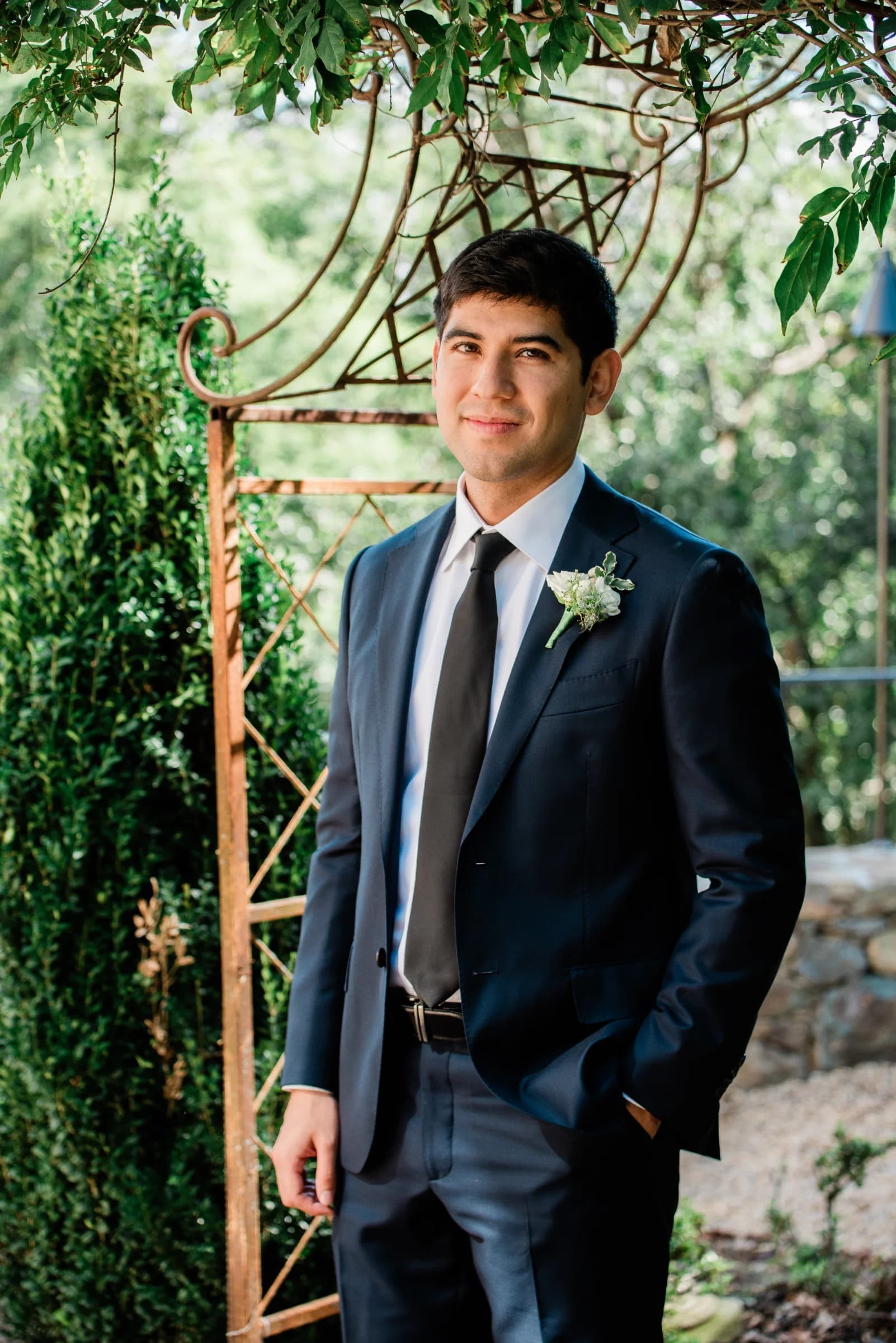 A man in a navy suit stands confidently under a leafy trellis, exuding elegance and charm in a serene outdoor setting.