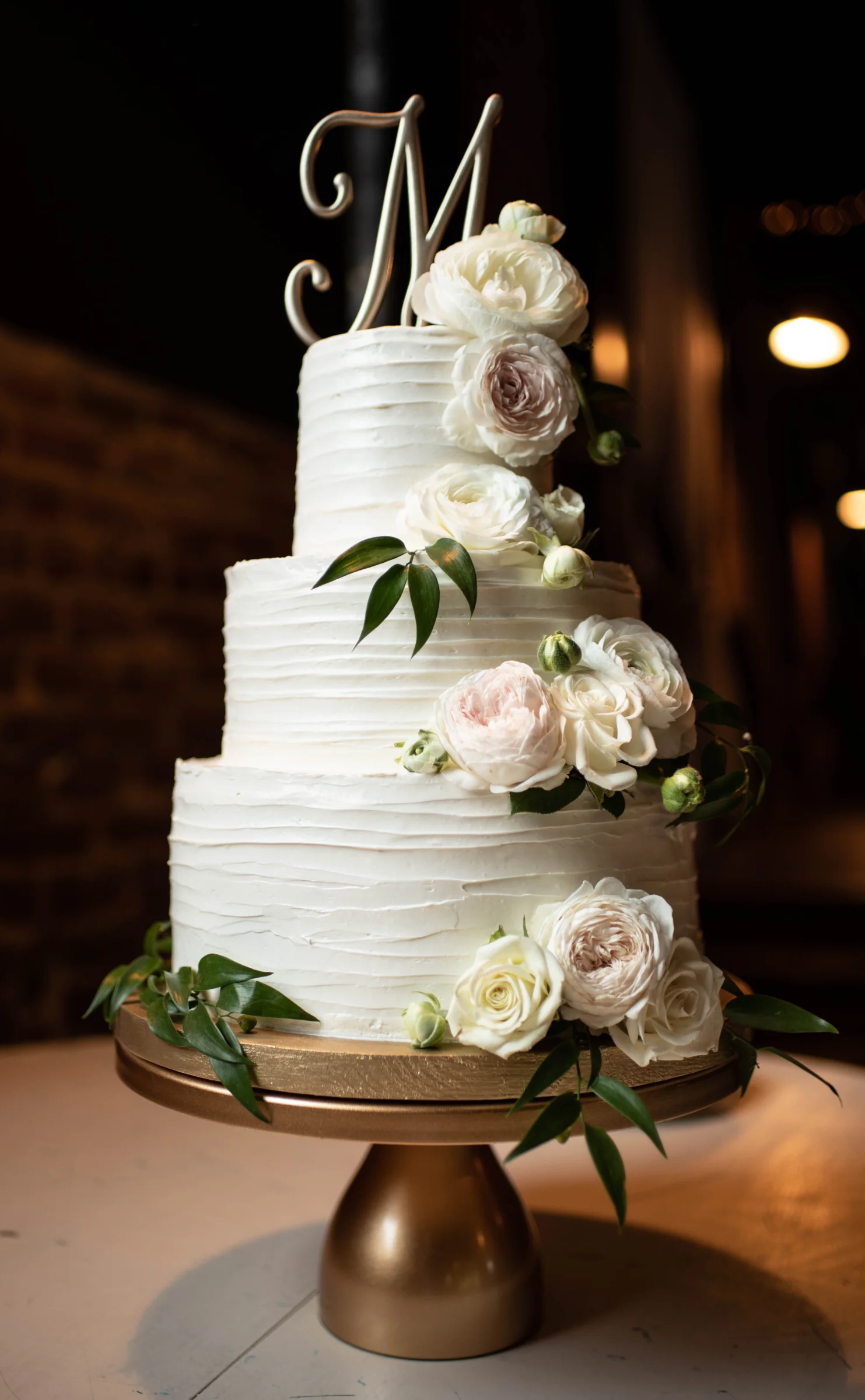 A three-tiered white cake adorned with soft pink and white flowers, topped with a silver "M" monogram, sits on a gold stand.