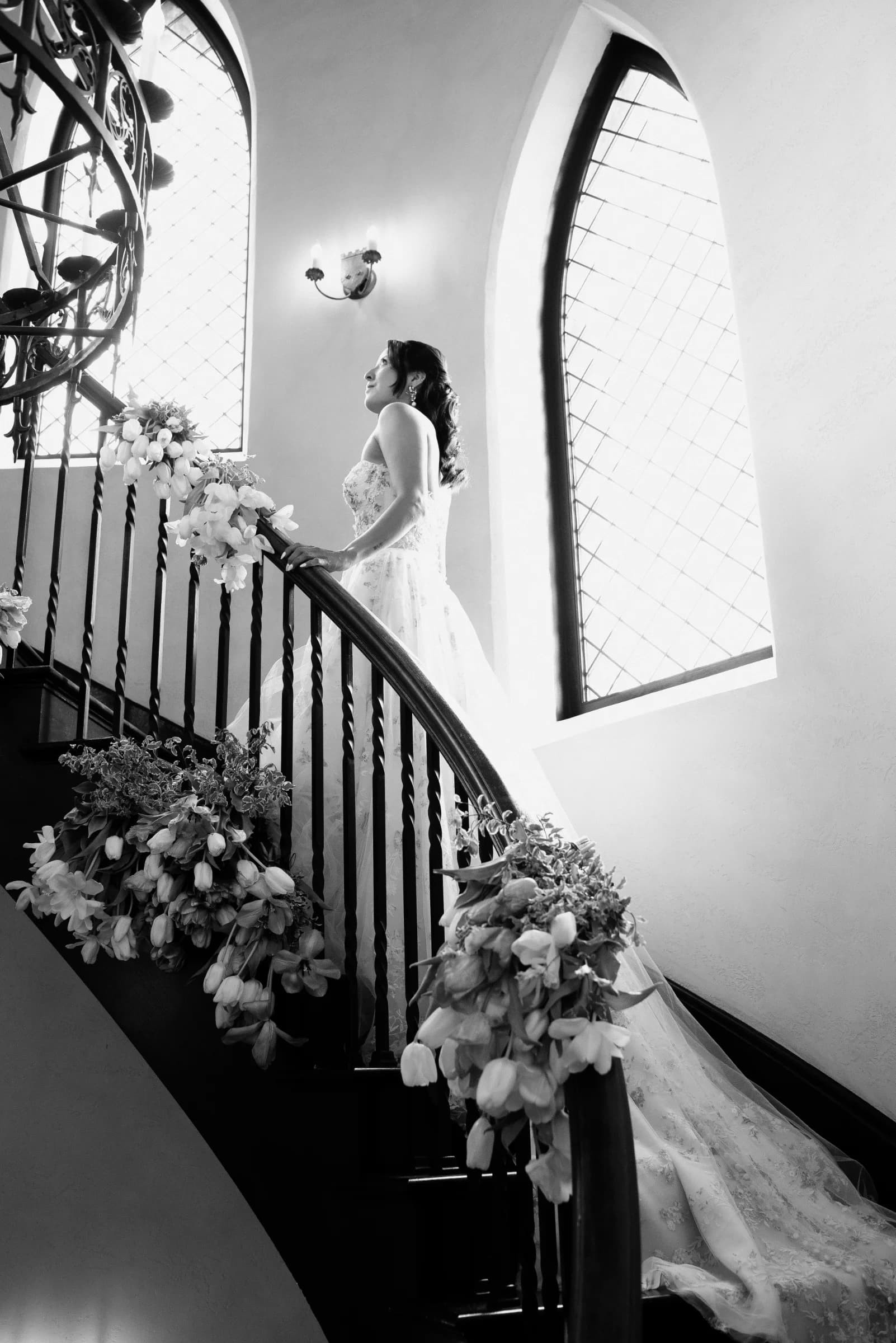 A bride in a flowing gown ascends a spiral staircase adorned with flowers, illuminated by soft light from arched windows.