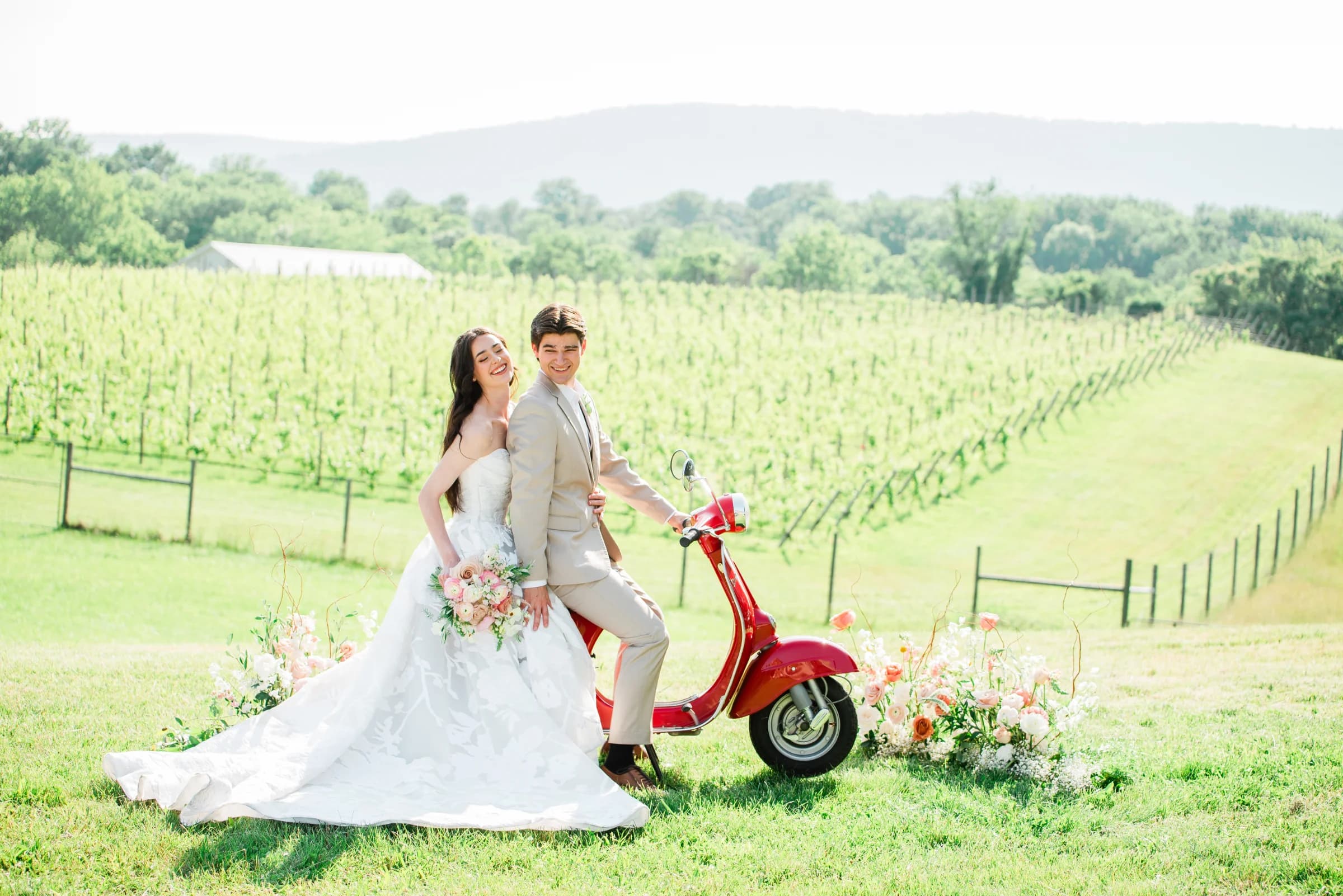 A joyful couple poses on a red scooter in a vineyard, surrounded by lush greenery and floral arrangements.