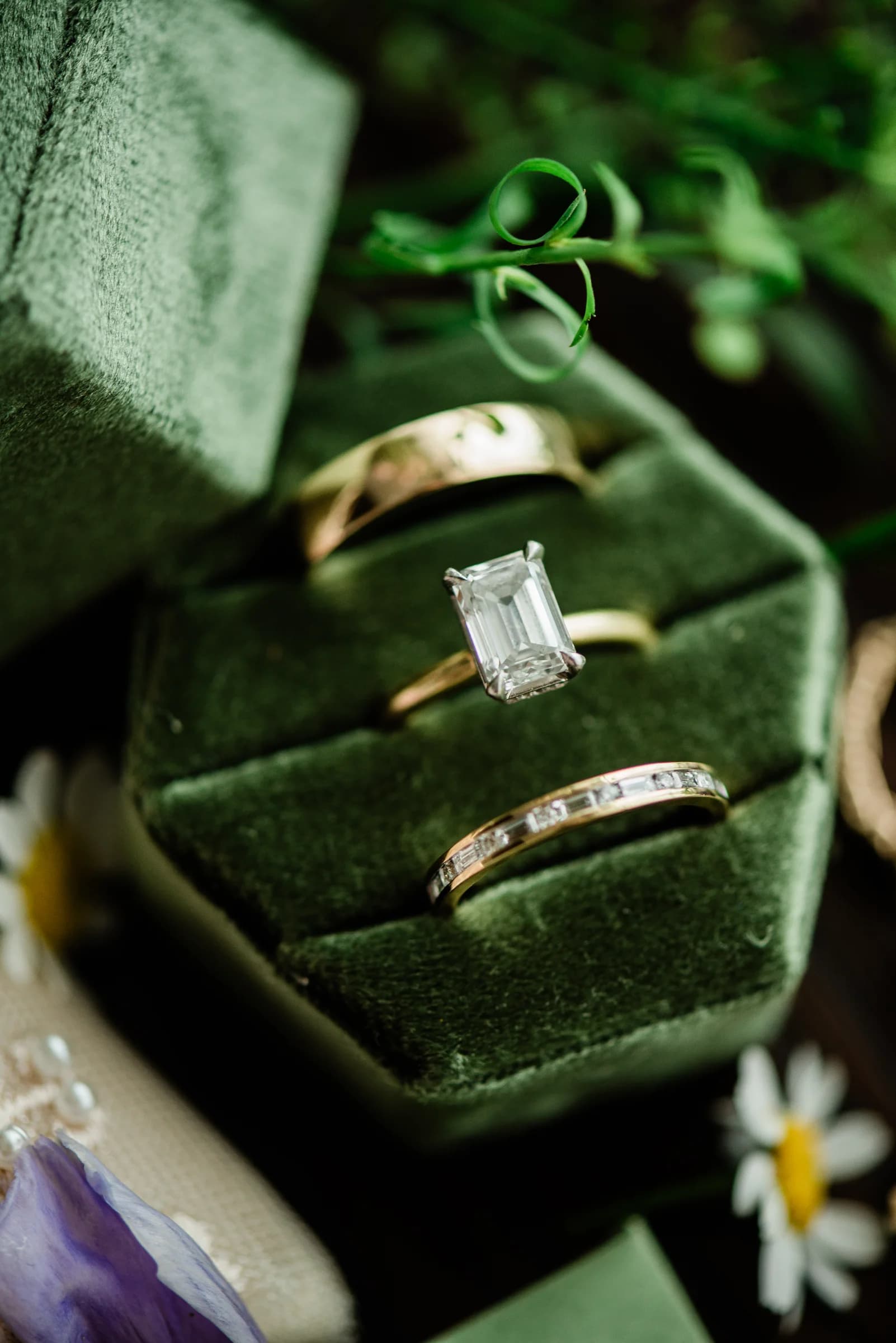 Three elegant rings, including a diamond solitaire, rest on a plush green display surrounded by delicate flowers.