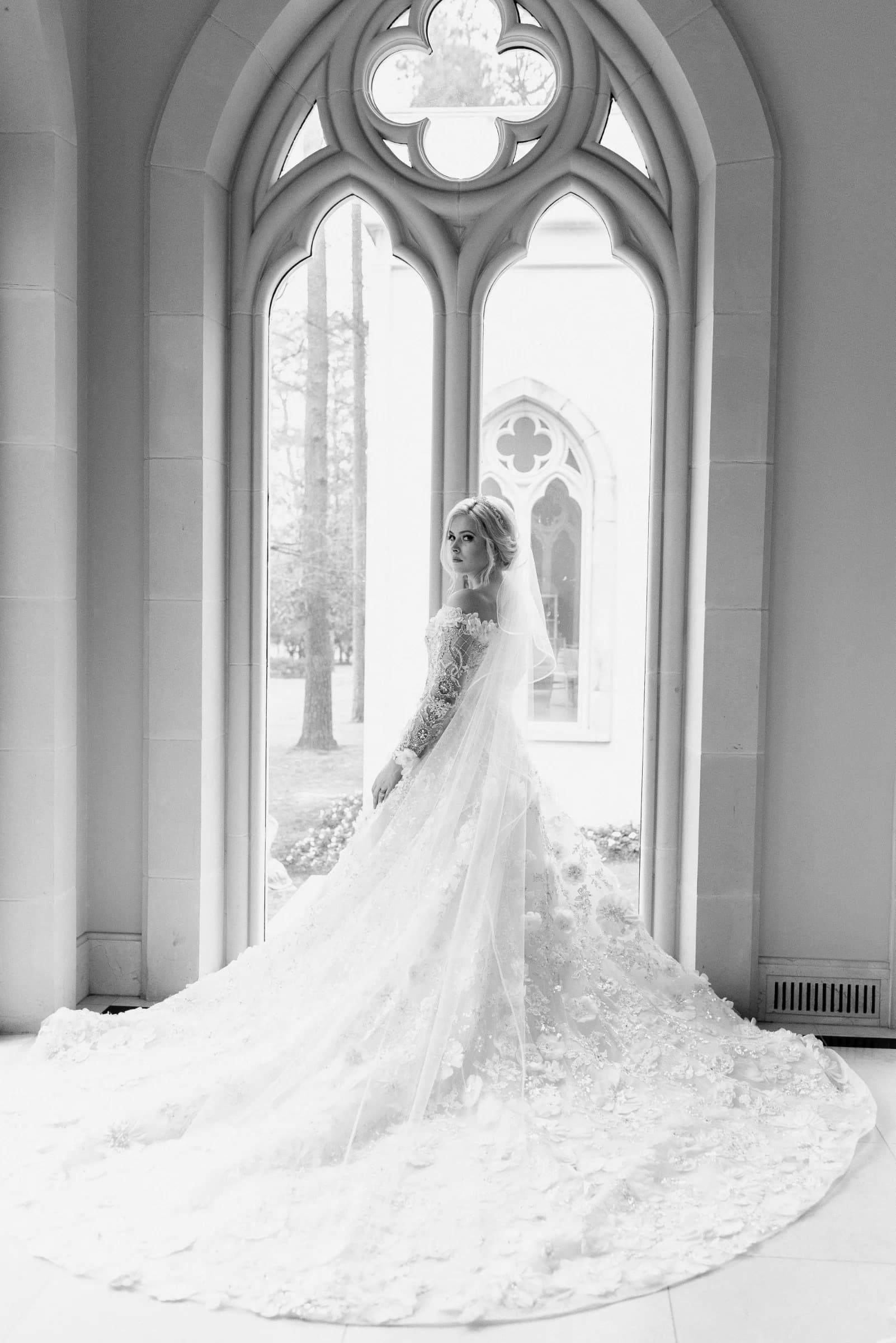 A bride in an intricate gown stands gracefully by a large arched window.