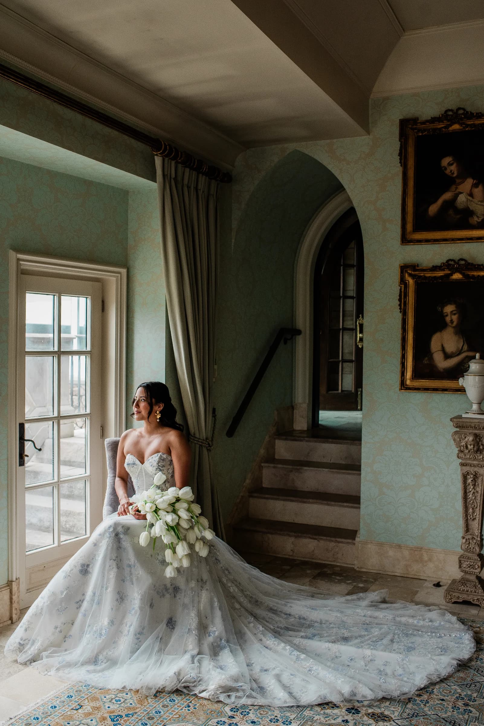 A bride in an intricately detailed gown stands in a softly lit grand hallway.